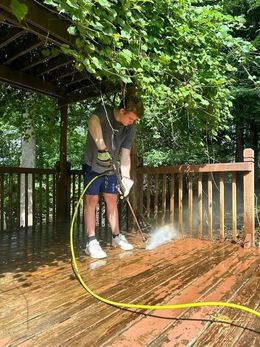 Person power washing a wooden deck with a yellow hose under a pergola.