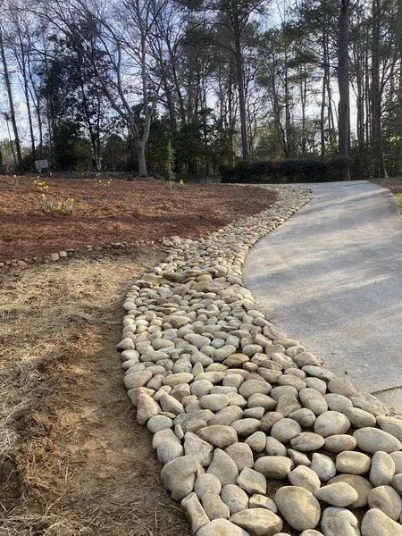 A gravel pathway alongside a concrete driveway, bordered by rounded rocks, in a wooded area.