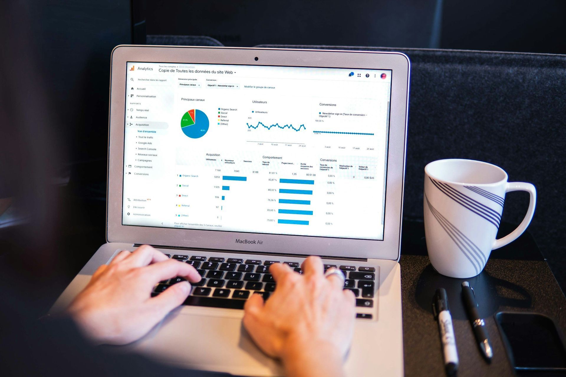 Person using a laptop displaying analytics data, with a coffee mug and pens on the side.