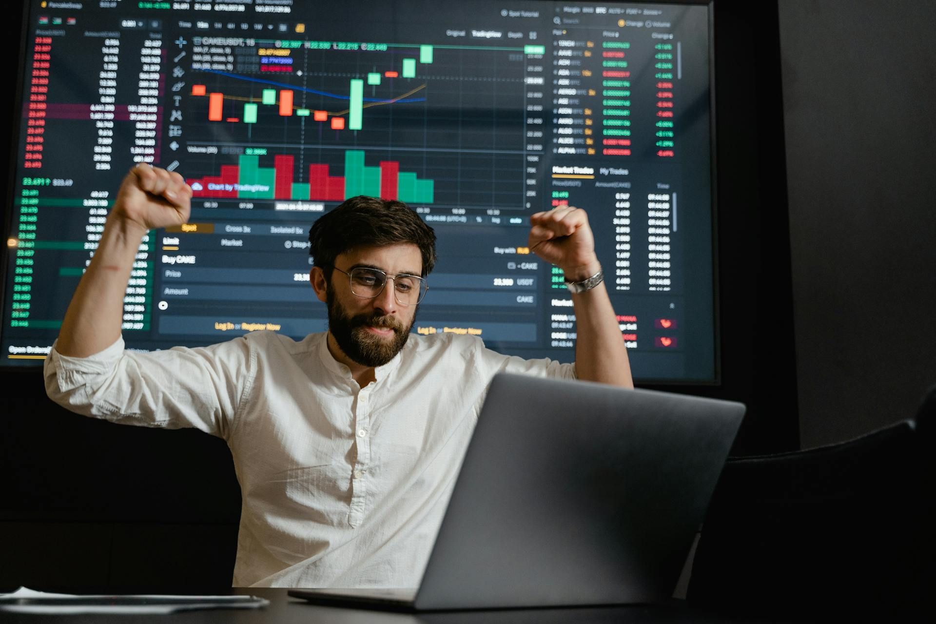 Man celebrating success in front of a laptop and stock market charts on a screen, raising his fists with joy.