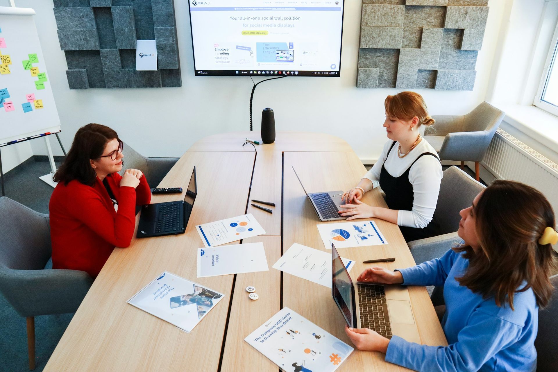 Three women are sitting at a table with laptops in a conference room.