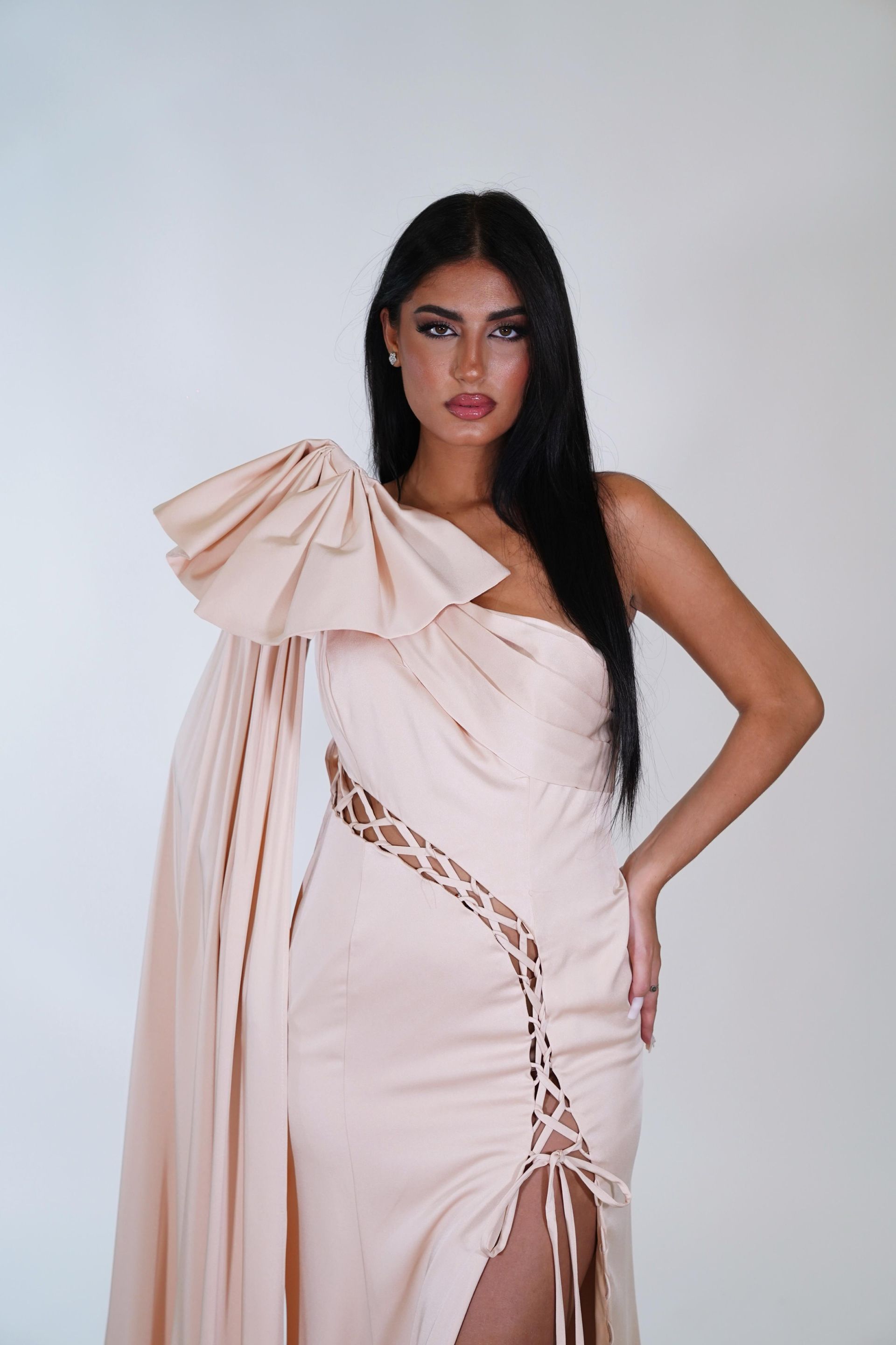 Una donna con un lungo abito bianco con un fiocco sulla spalla è in piedi davanti a un muro bianco.