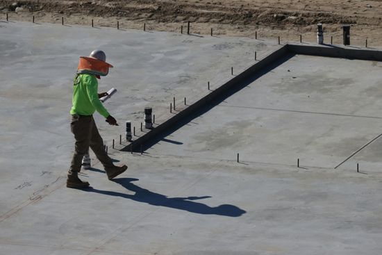 A construction worker in a high-visibility lime shirt walks across a flat concrete foundation at a construction site.