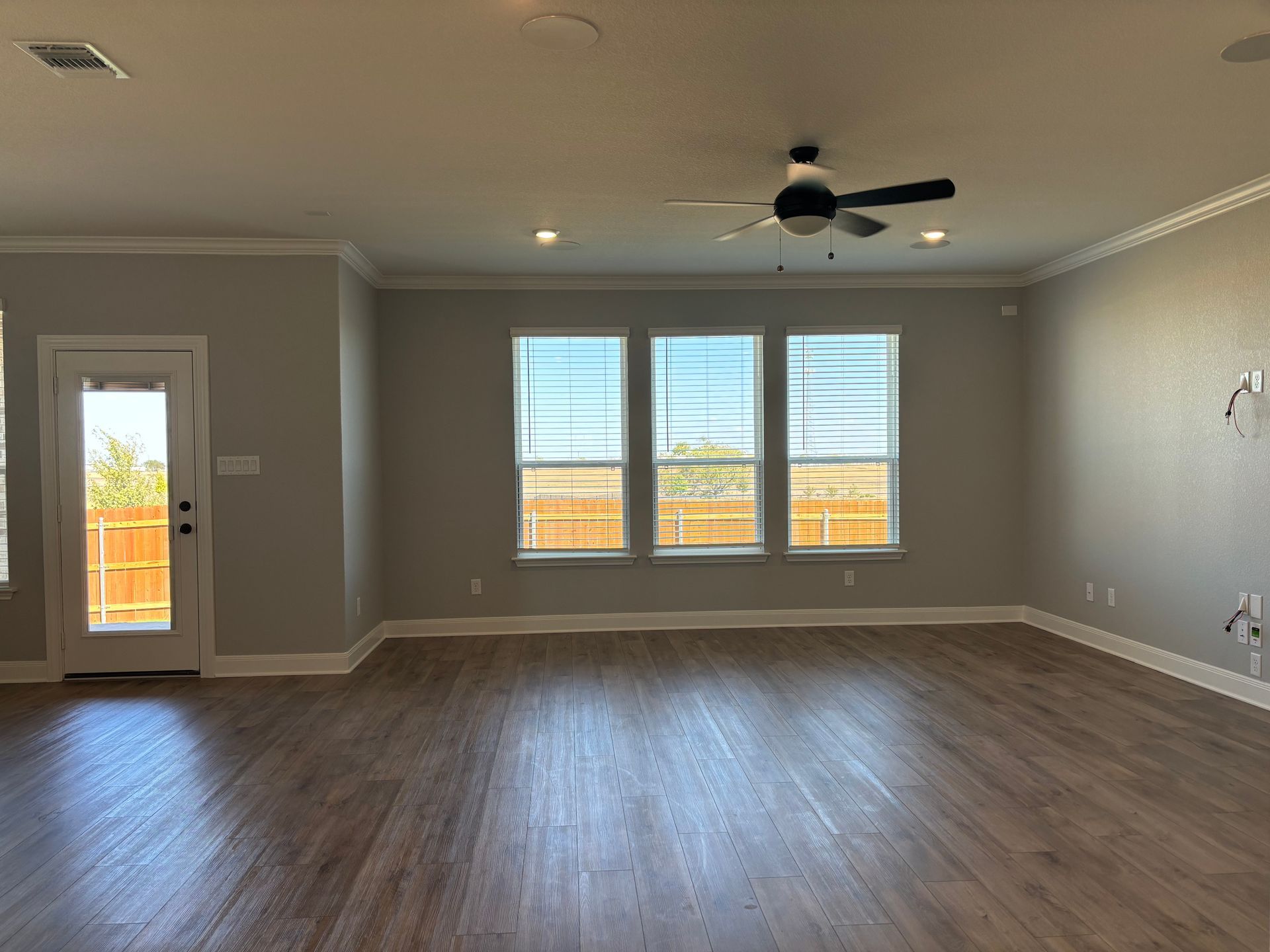 Empty modern living room with wood floors, gray walls, ceiling fan, and three windows.