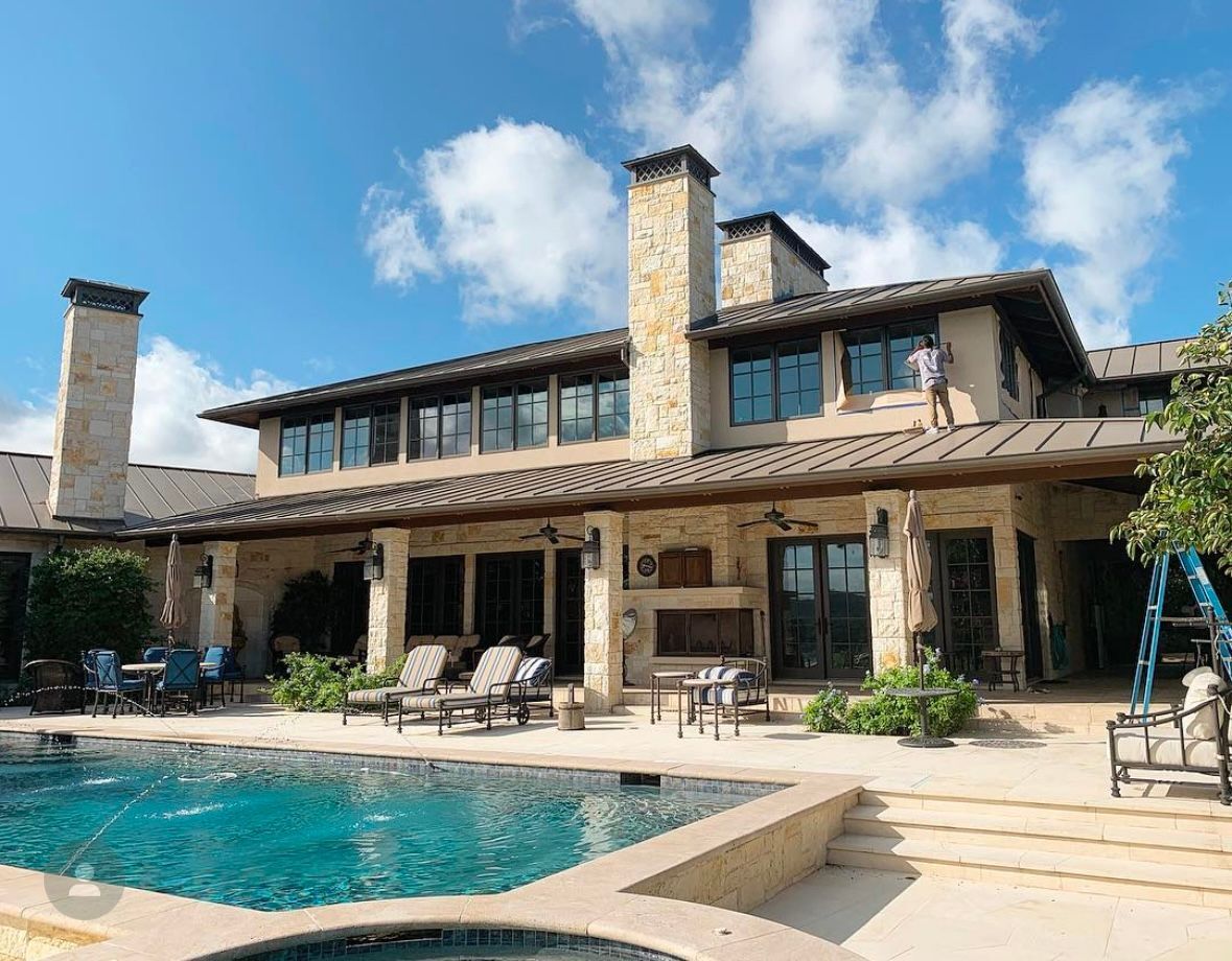 Large, two-story house with a pool, patio, and stone chimney on a sunny day. A person is working on the roof.