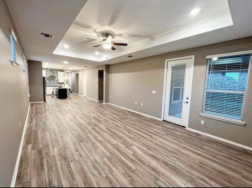 Empty open-plan living area with wood-look flooring, tray ceiling, ceiling fan, and window blinds.