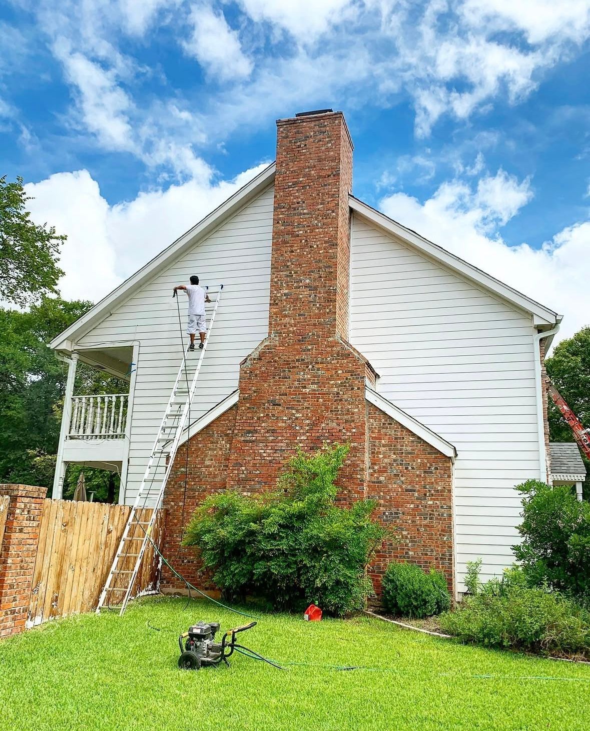 Person painting white siding on a house. Brick chimney, green lawn, ladder, and lawnmower in view.