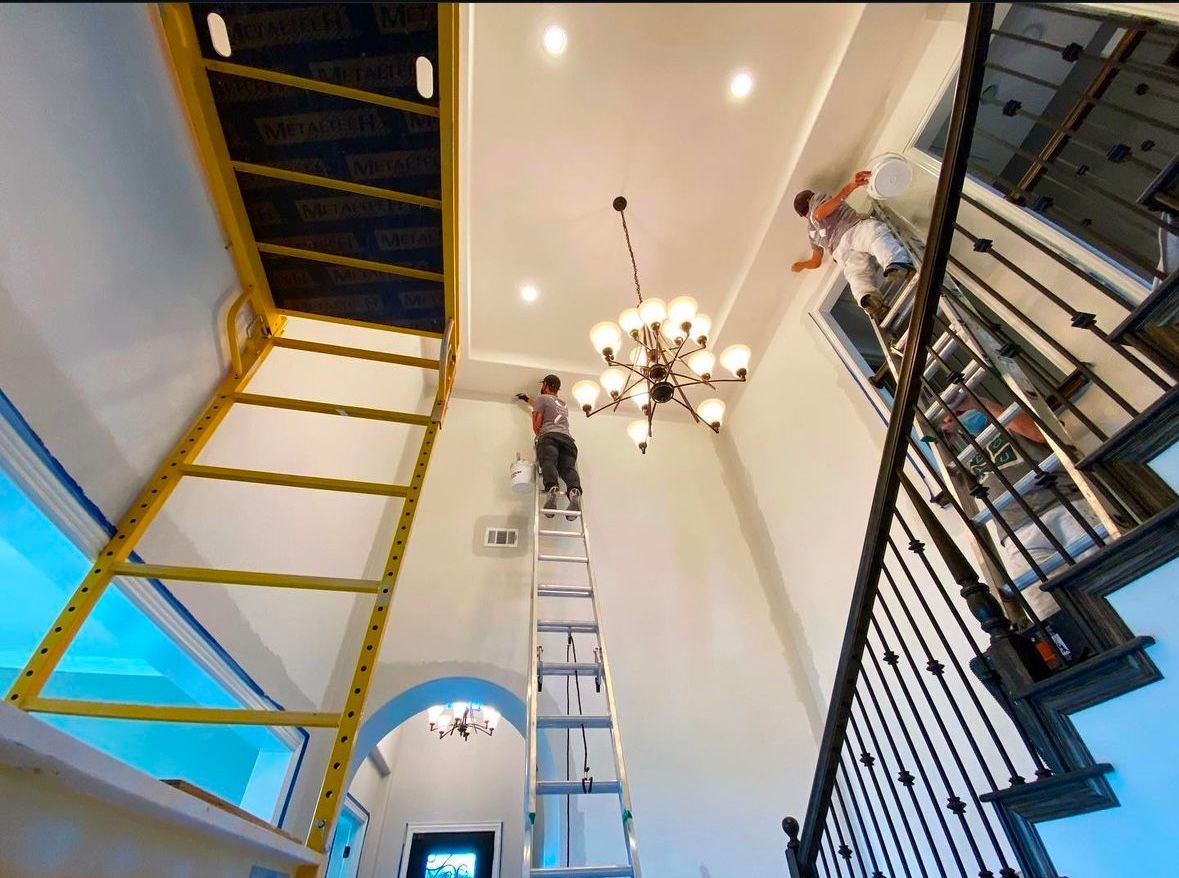 Two painters on ladders, working on a high ceiling in a well-lit interior space.