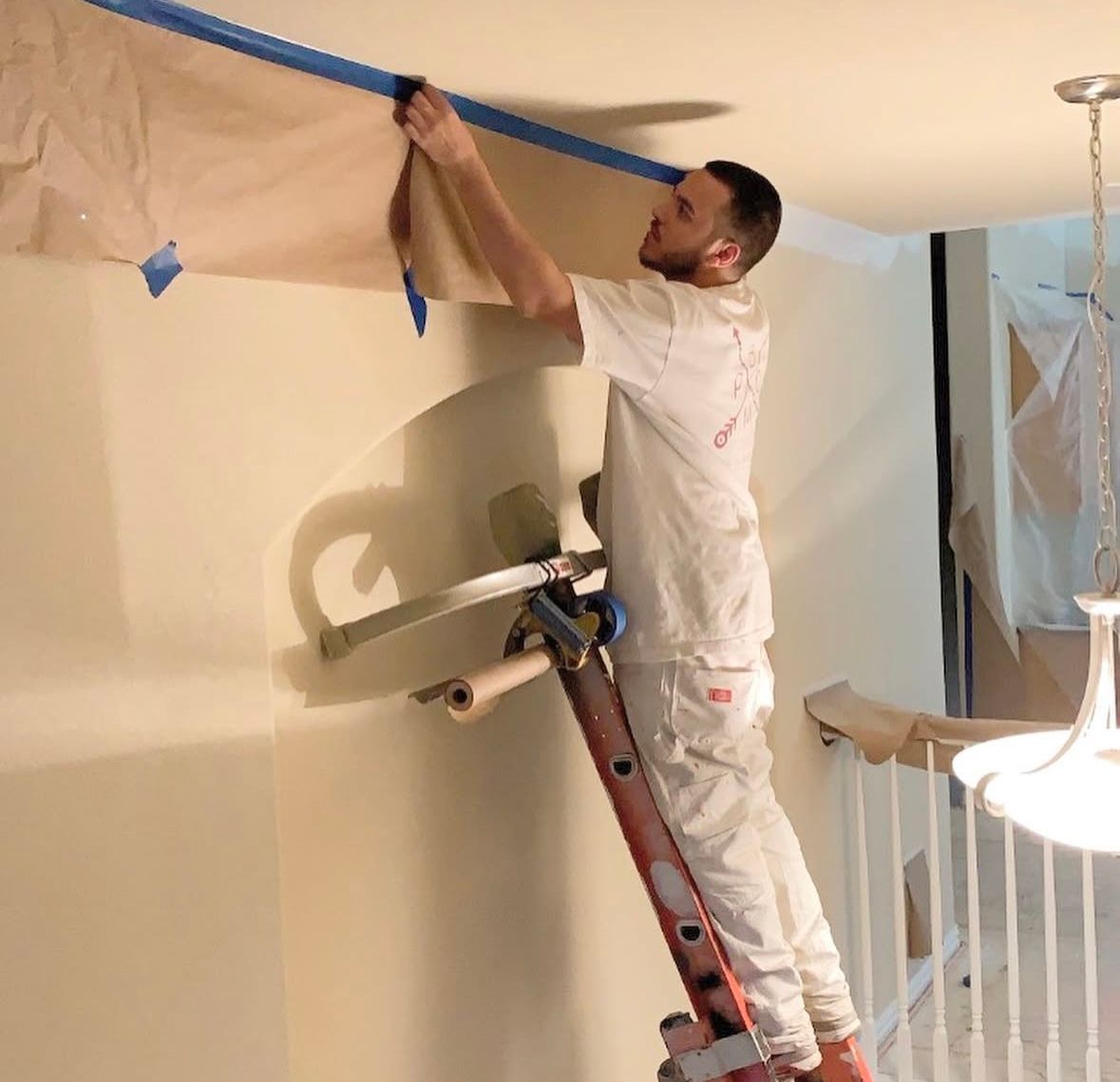 Painter taping a room ceiling while standing on a ladder, with plastic-covered furniture nearby.