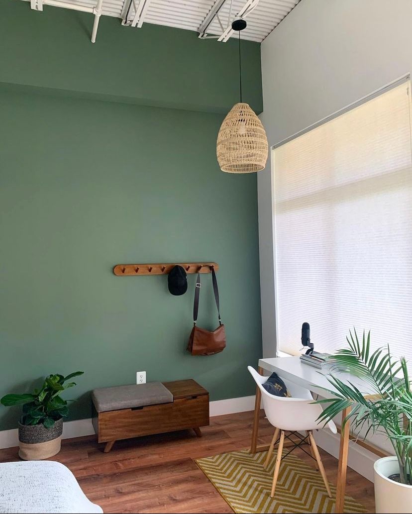 A bedroom with green walls , a desk , a chair , a wicker lamp , and a window.