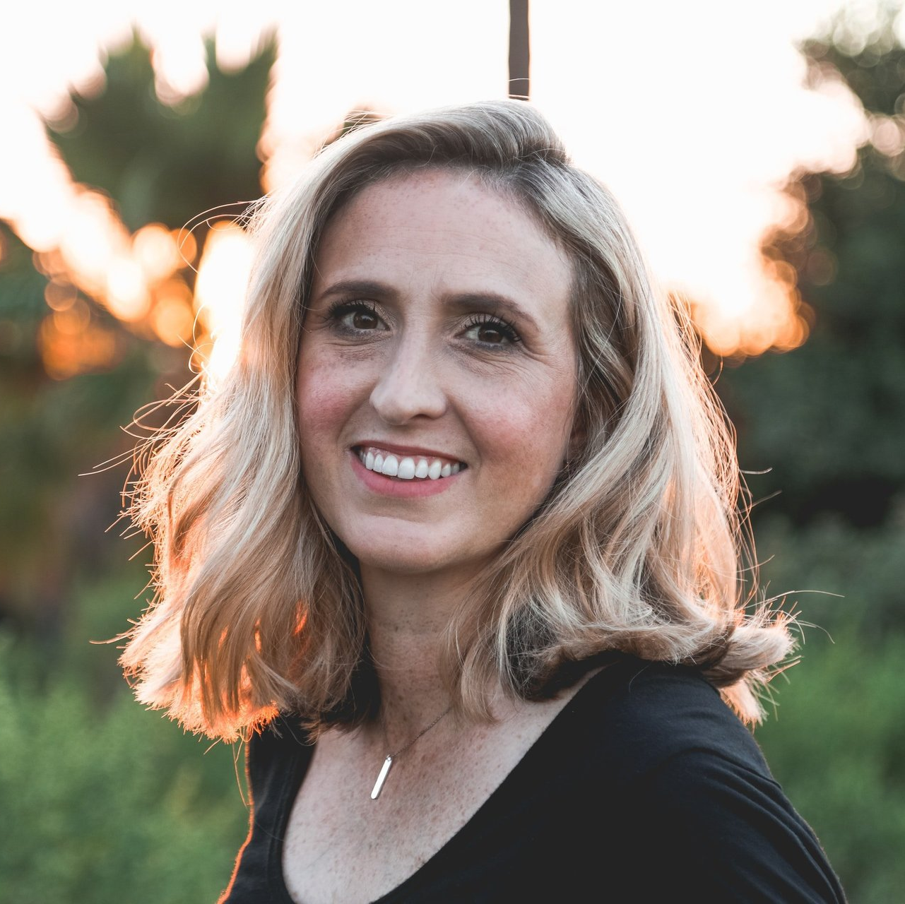 Woman with shoulder-length blonde hair smiles outdoors at sunset, wearing a black top and silver necklace.
