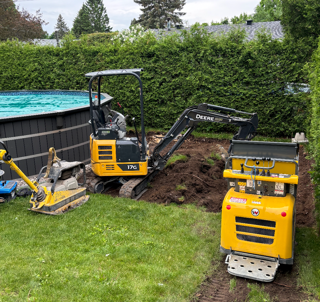 Une excavatrice jaune creuse un trou dans une cour arrière à côté d'une piscine.