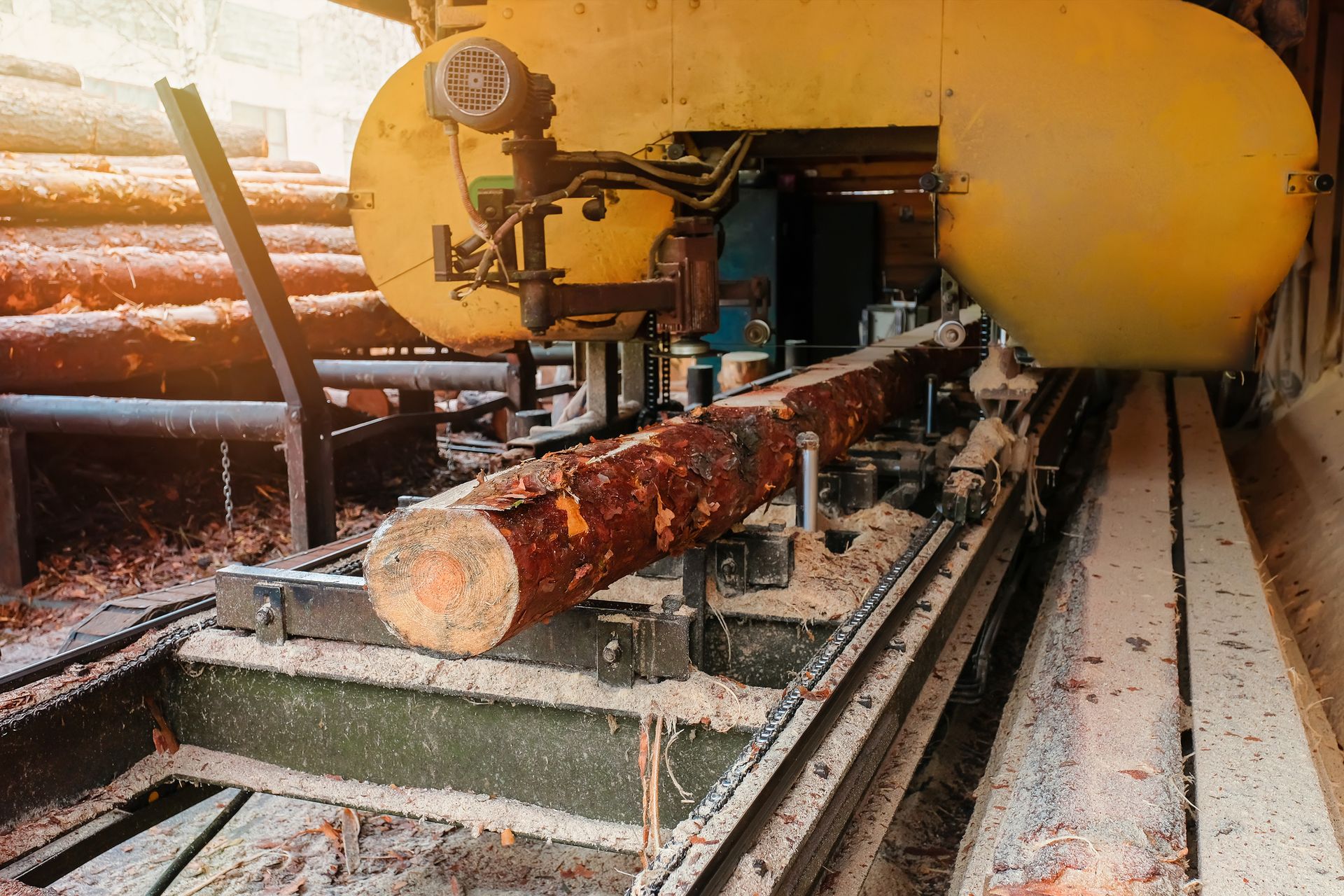 Sawmill machine cutting logs into lumber with stacked logs in background.