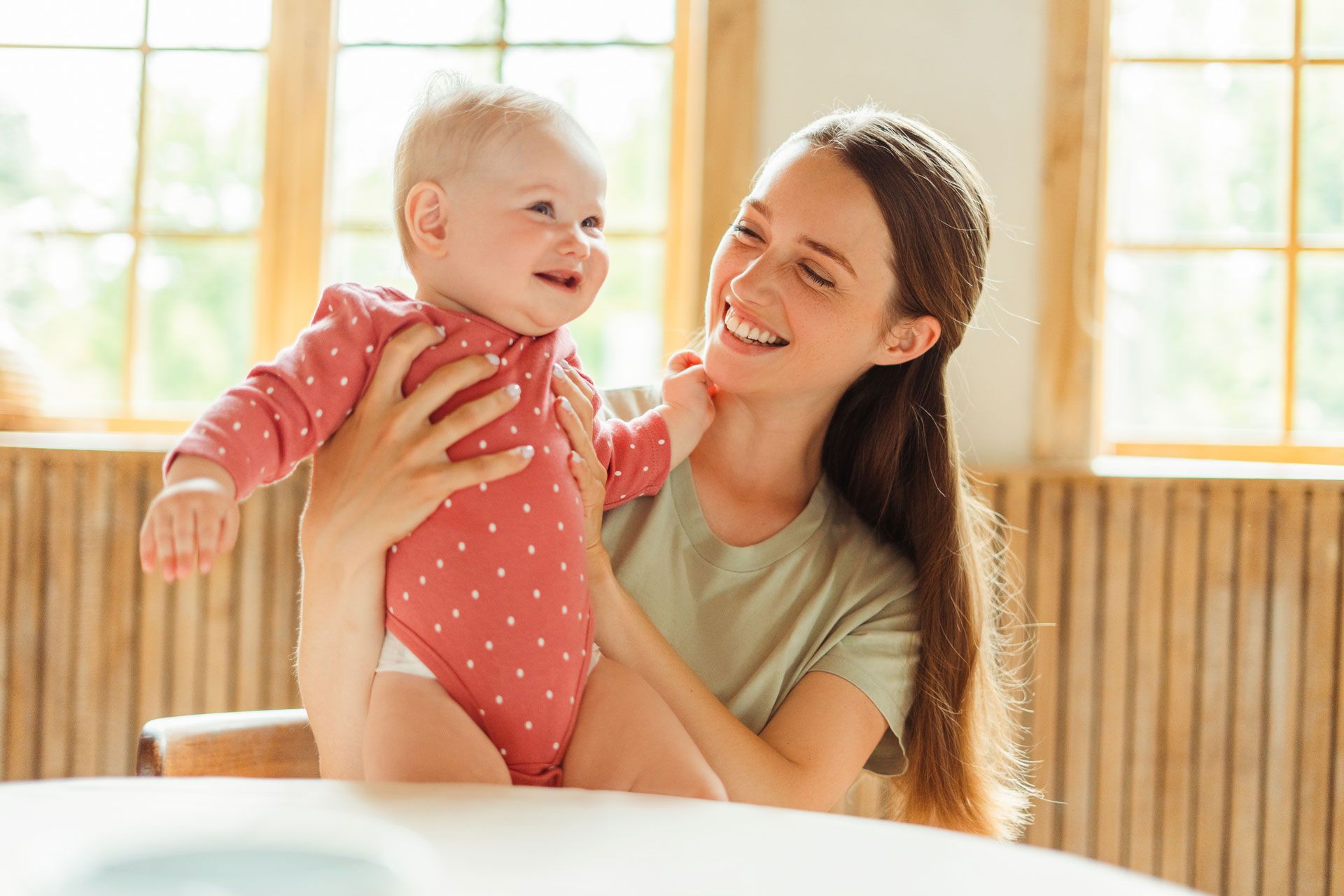 Woman holding a smiling baby indoors, both looking at each other. Soft natural light, warm colors.