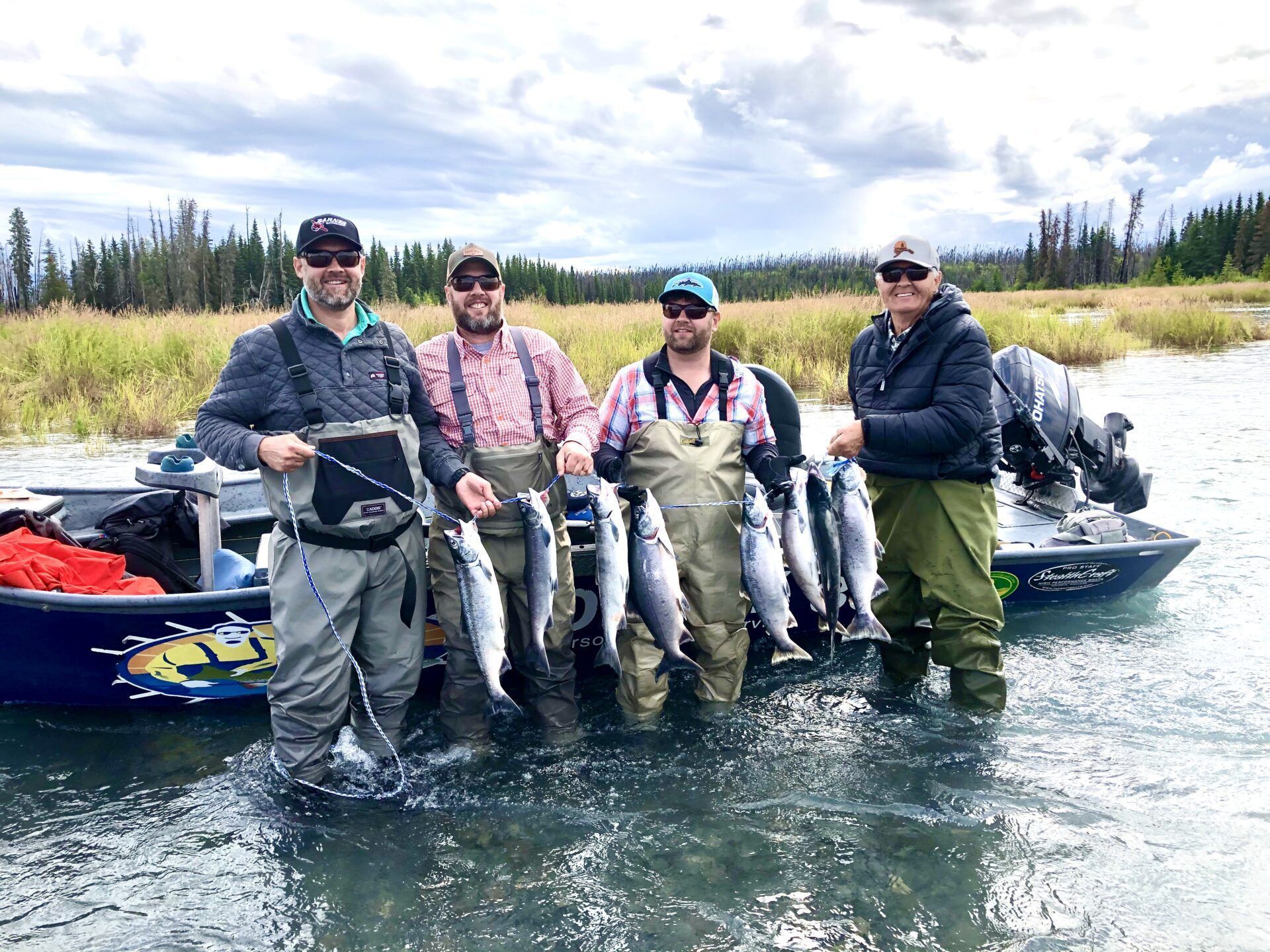 Four people in waders standing in a river by a boat, each holding a fish they have caught.