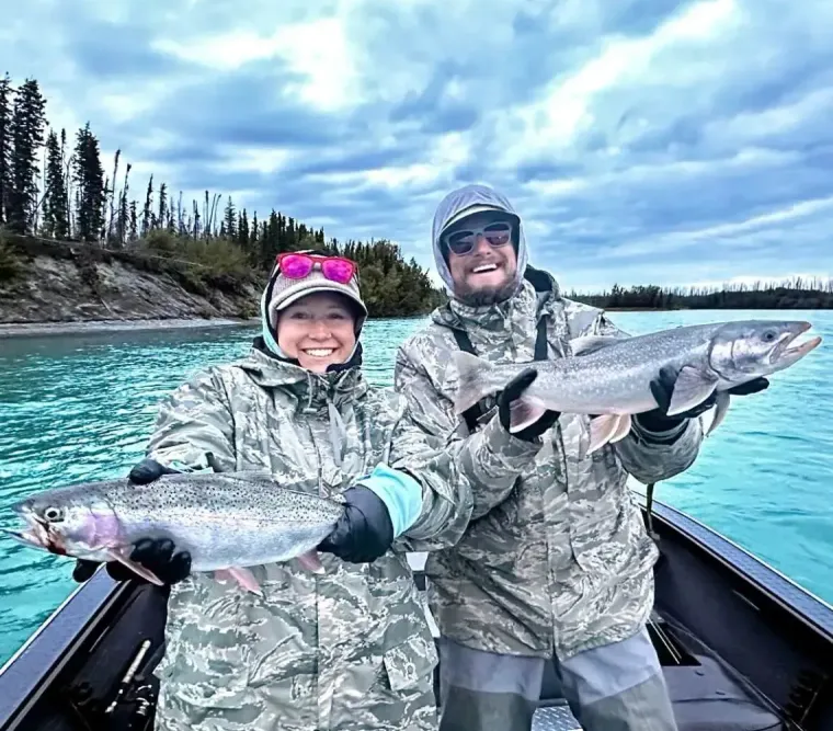 Two people in camouflage fishing gear hold trout while standing in a boat on a blue river under a cloudy sky.