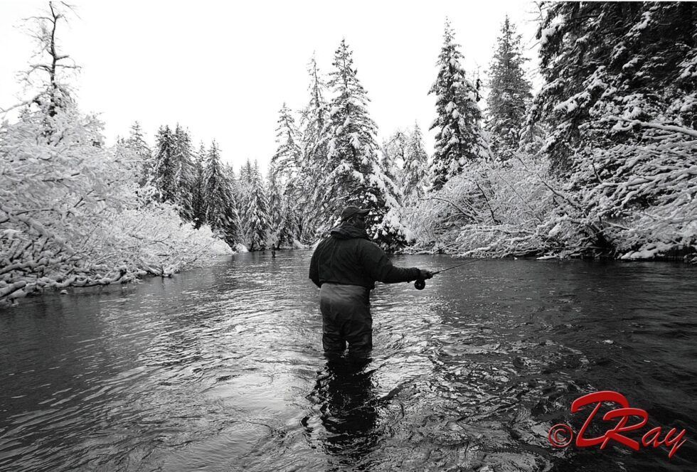 A person in dark fishing gear stands in a river surrounded by snow-covered trees in a black-and-white scene.