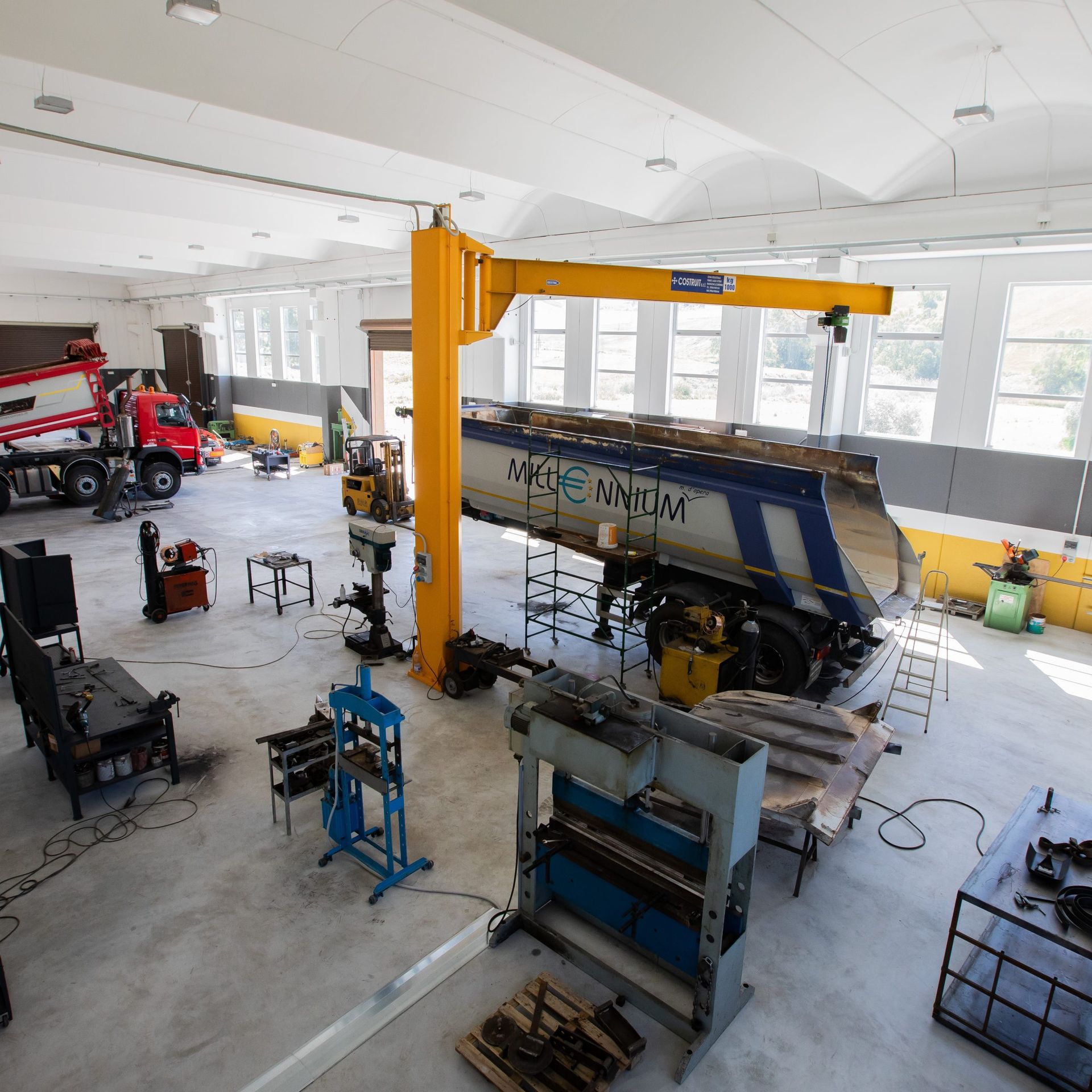 Interno di un'officina con macchinari e un grande rimorchio per camion in riparazione. Gru e attrezzature gialle.