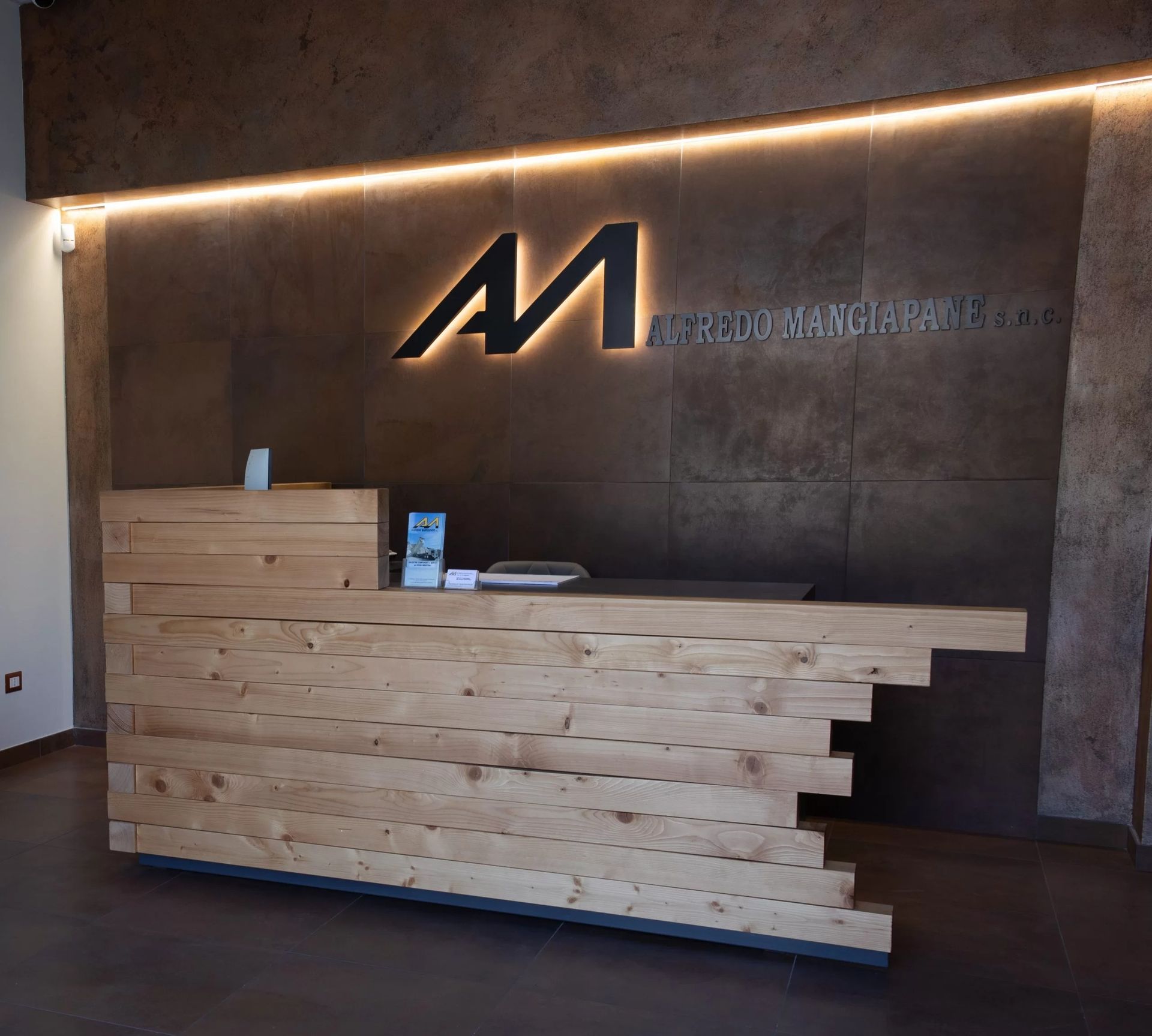 Bancone della reception in legno con il logo aziendale su una parete dalla texture scura, illuminato dall'alto.