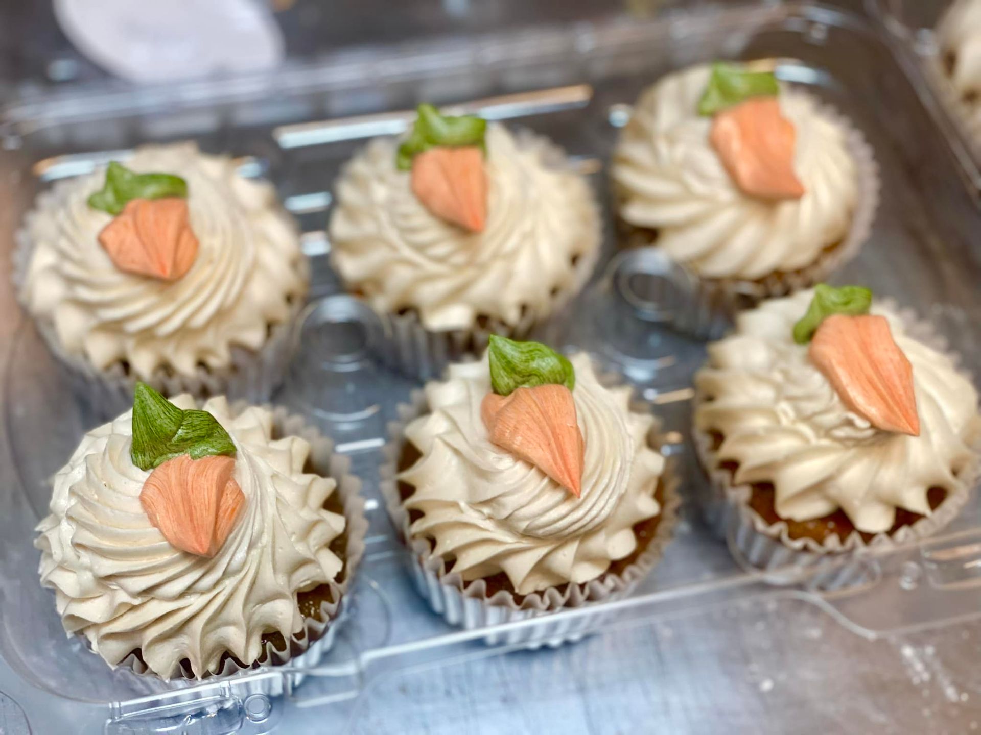 Six carrot cupcakes in a clear container, each topped with swirled tan frosting and a carrot-shaped decoration.