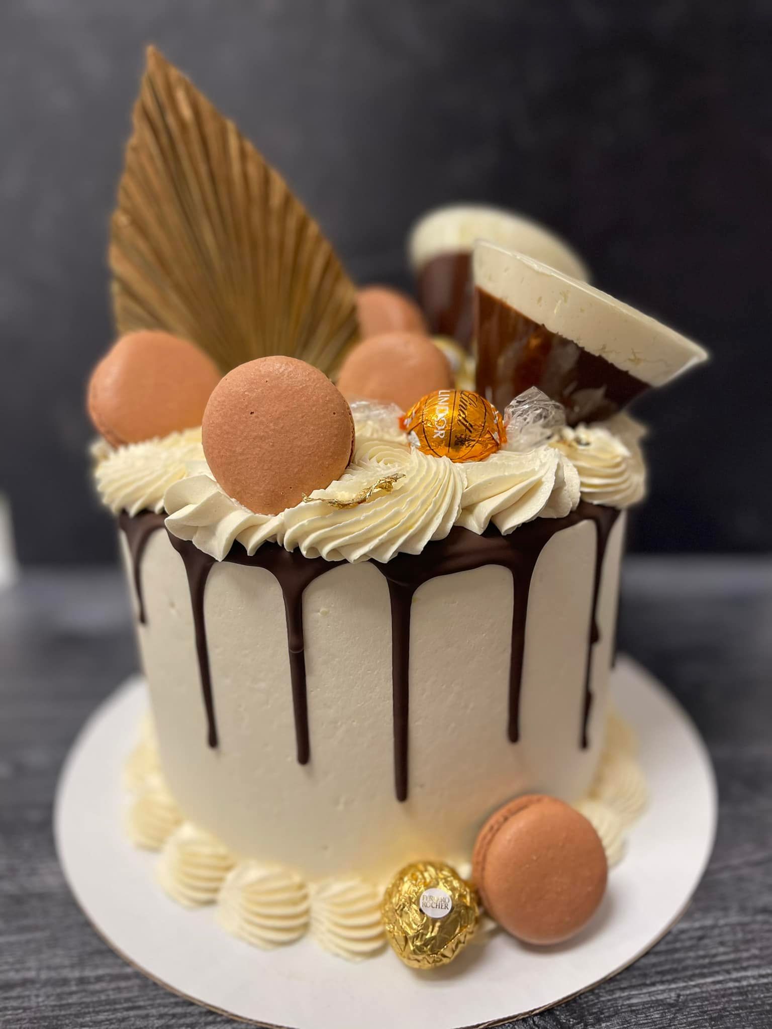 Cake with white frosting, chocolate drip, macarons, and decorative elements.