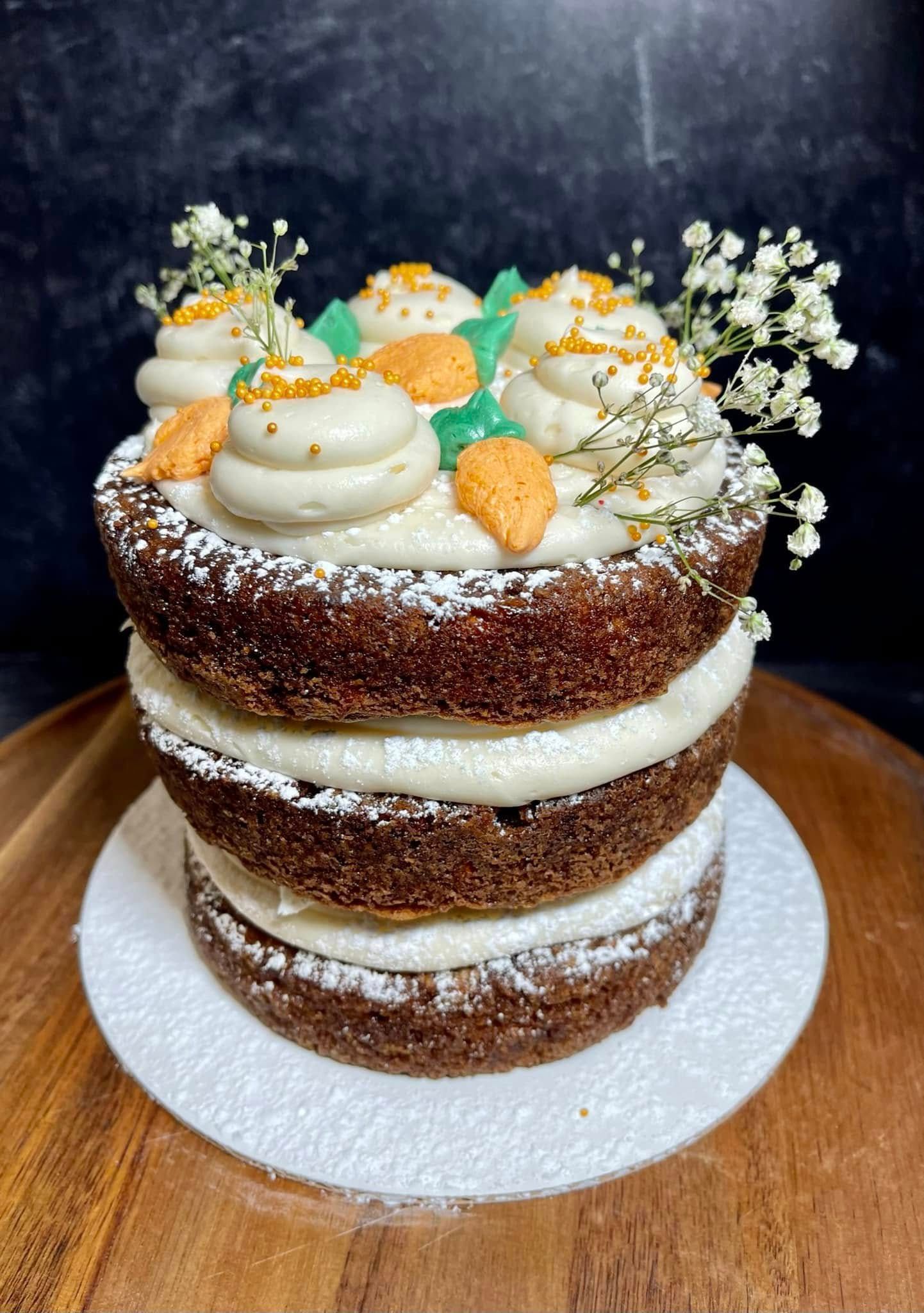 Layered carrot cake with cream cheese frosting, decorated with sugared carrots and flowers on a white plate.