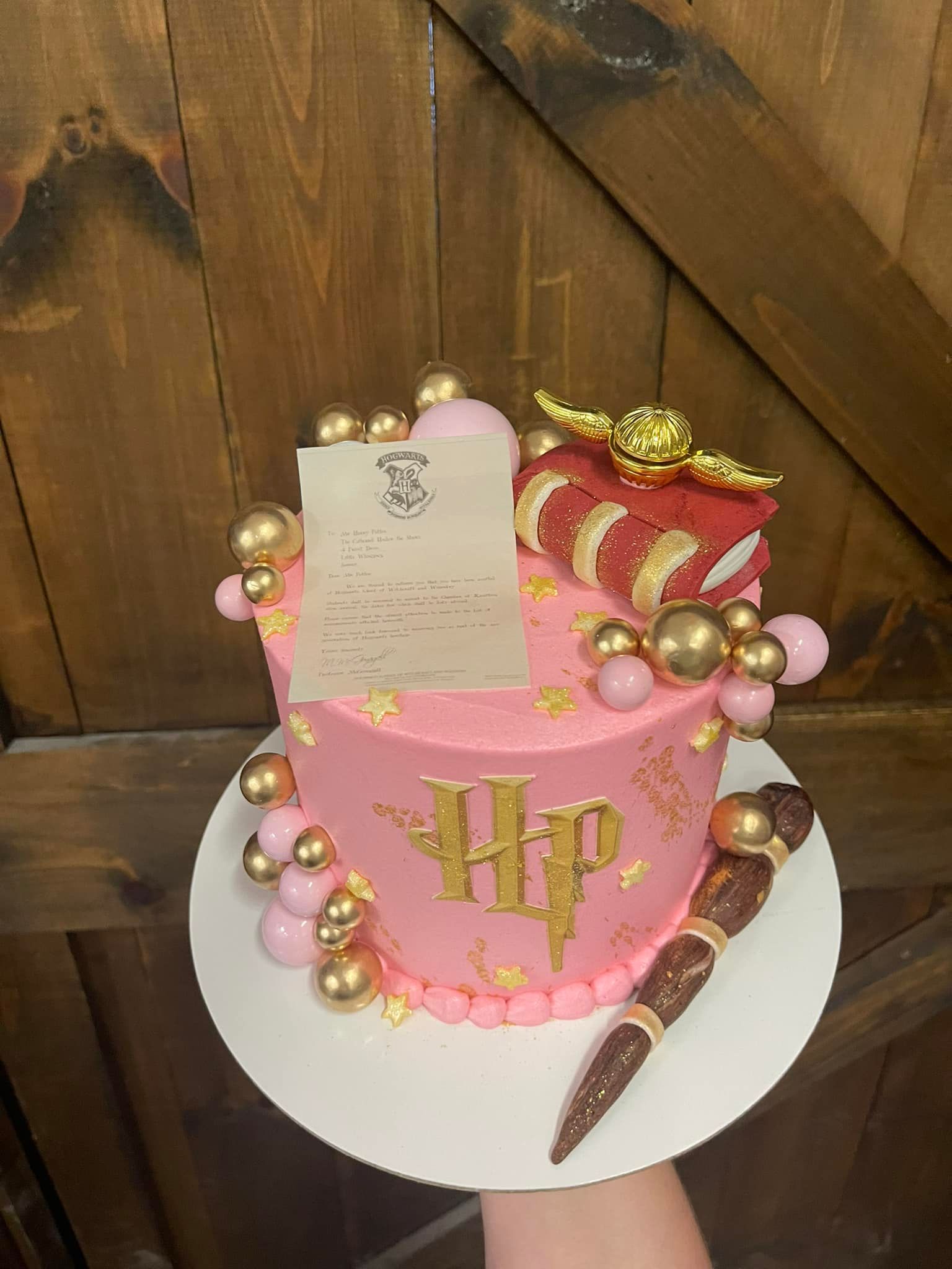 Pink Harry Potter-themed cake with gold decorations, wand, and letter scroll on a wooden background.