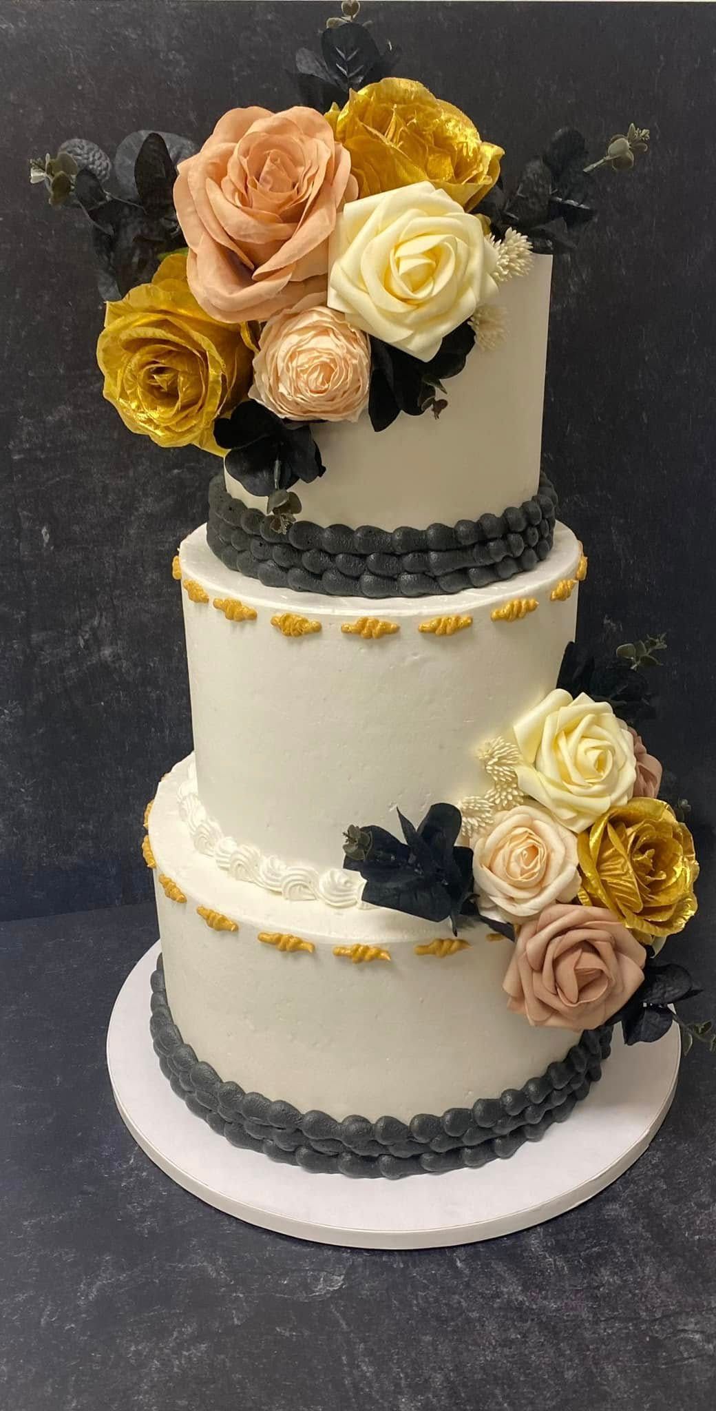 Three-tiered white cake, decorated with dark grey trim, gold accents, and colorful rose flower arrangements.