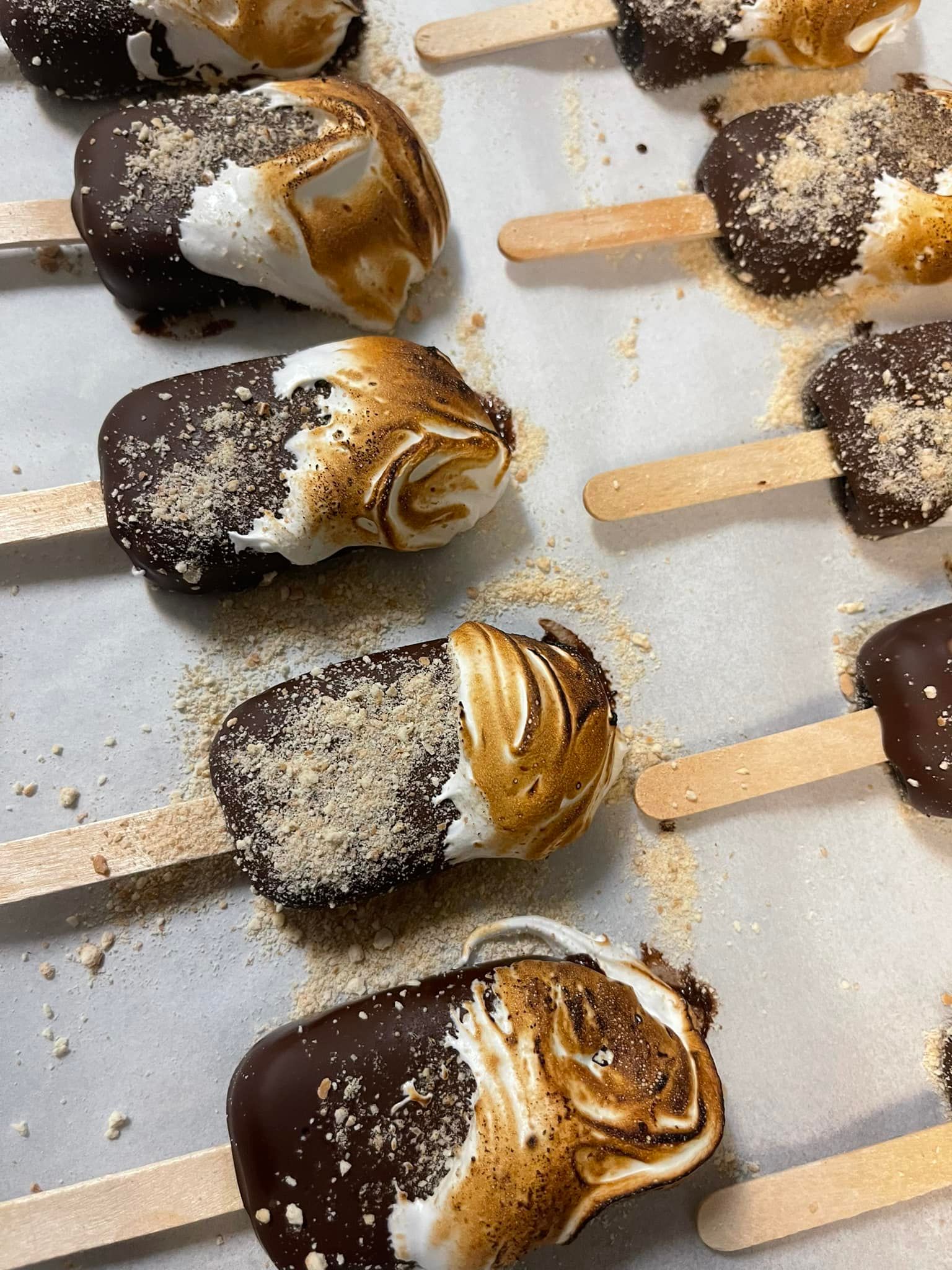 S'mores on sticks: chocolate-covered, marshmallow-topped treats with graham cracker crumbs, on a baking sheet.