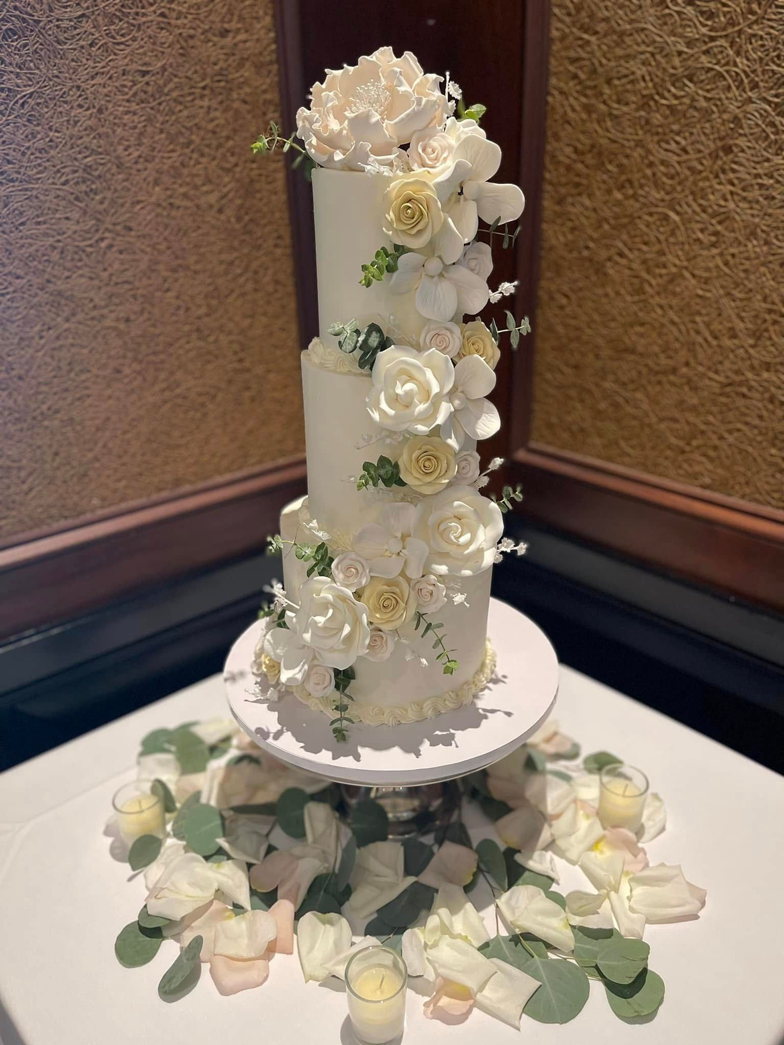 Three-tiered white cake with floral decorations on a pedestal, surrounded by petals and greenery.