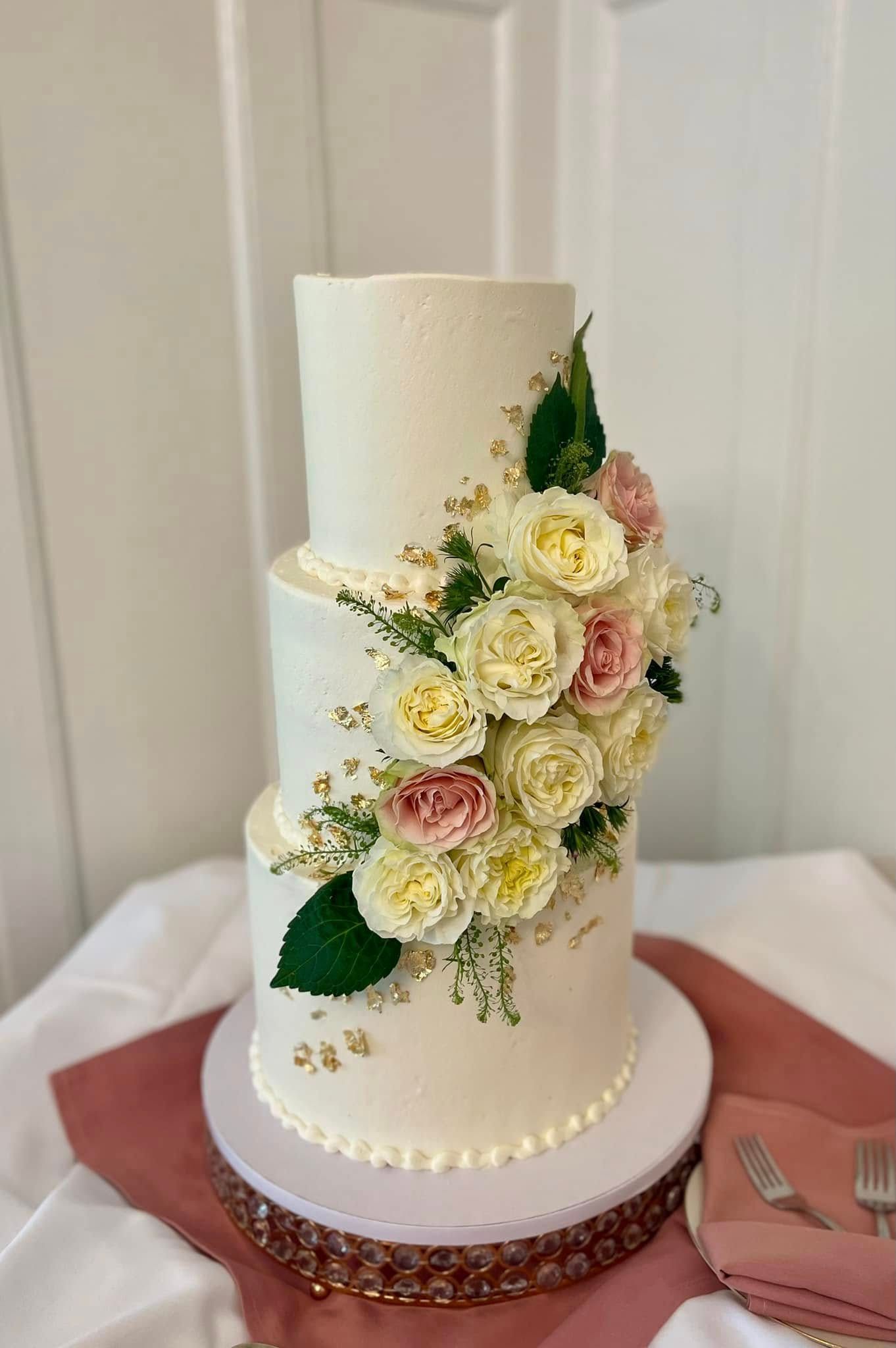 Three-tiered white cake, decorated with flowers and greenery, sitting on a table with a pink tablecloth.