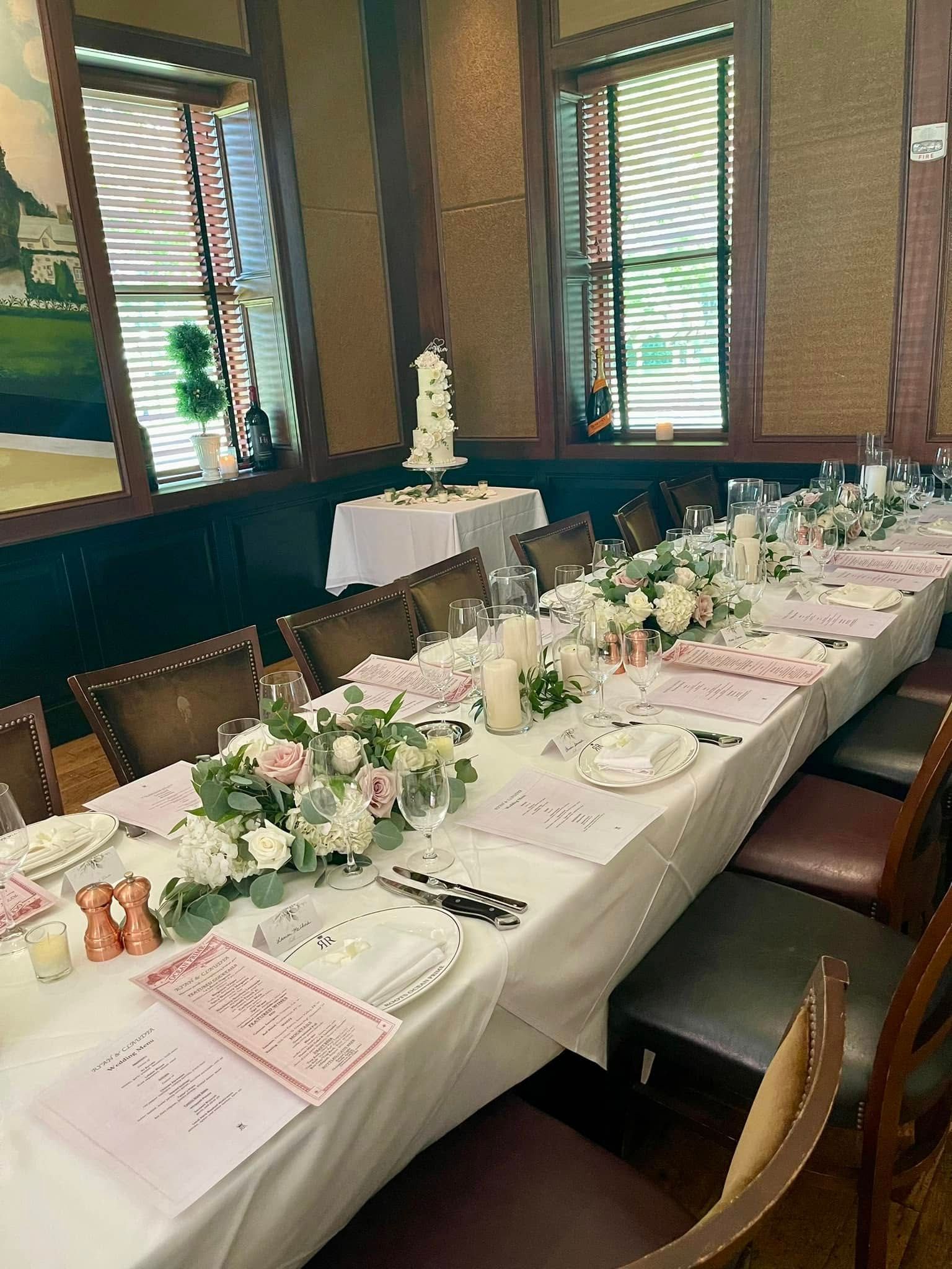 Long wedding reception table set with floral arrangements and a cake in a restaurant.