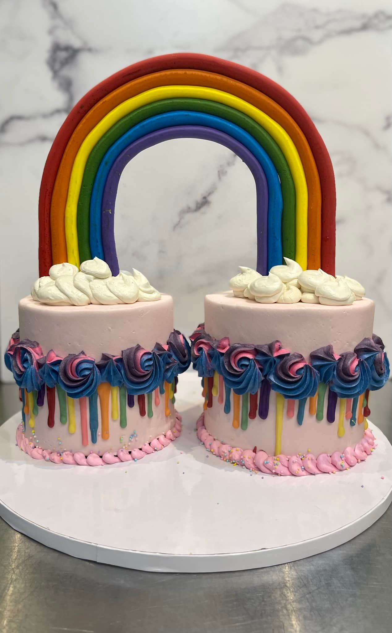 Two pink cakes with rainbow drip, clouds, and a rainbow arch.