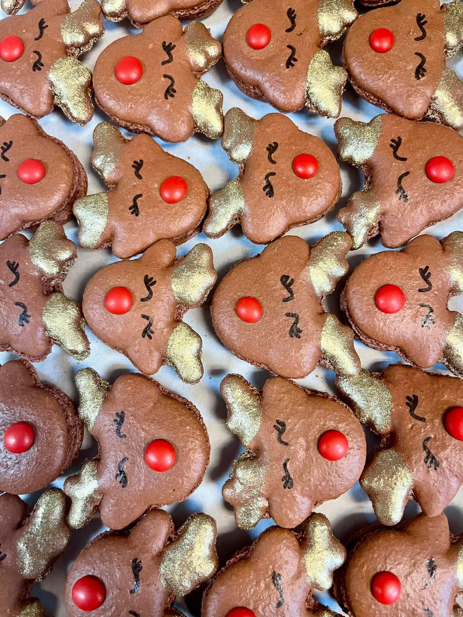 Rows of reindeer-shaped chocolate macarons with red noses and golden antlers, arranged on a surface.
