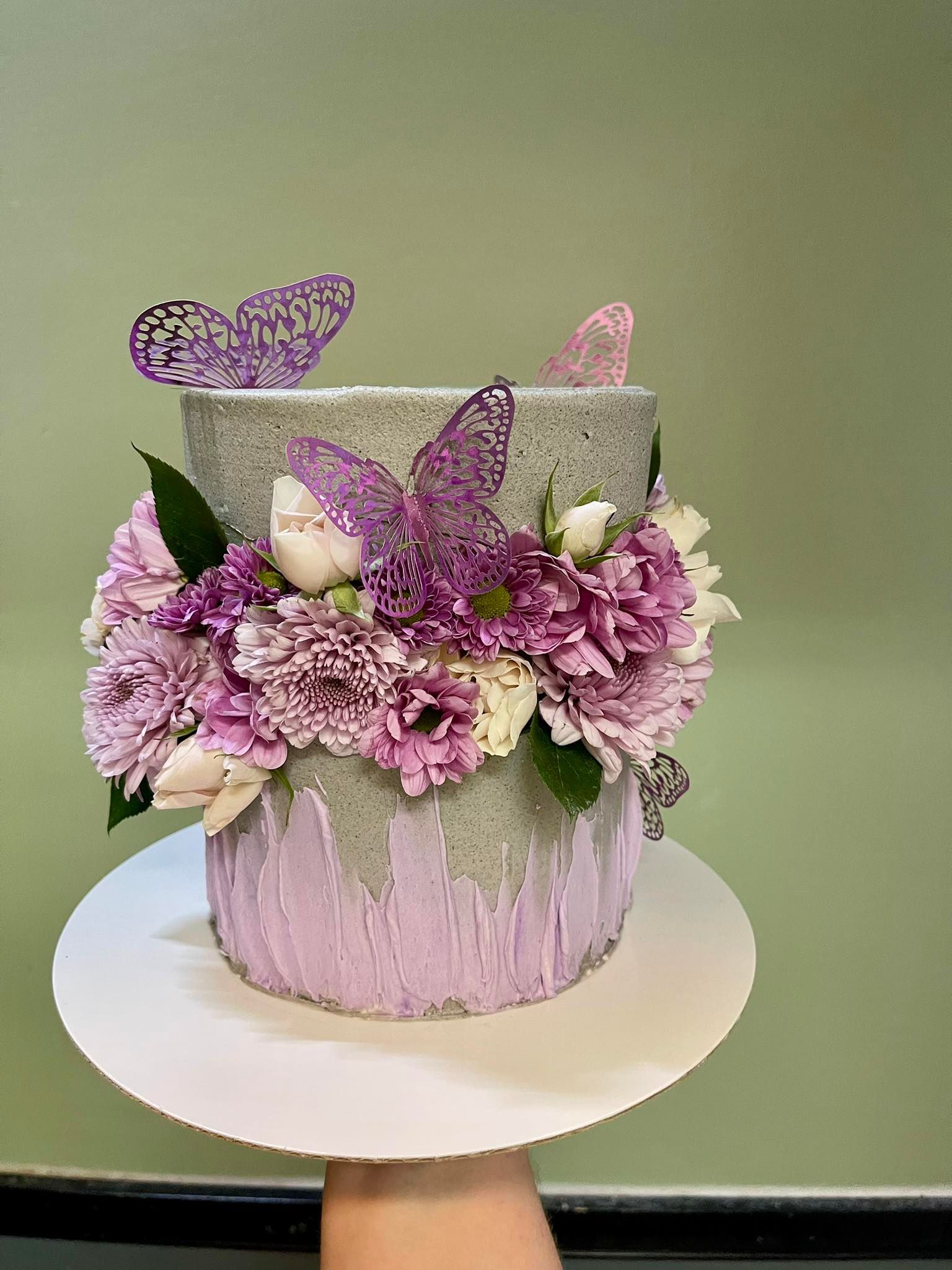Two-tiered cake with gray frosting, purple drizzles, and flowers, adorned with purple butterfly decorations.