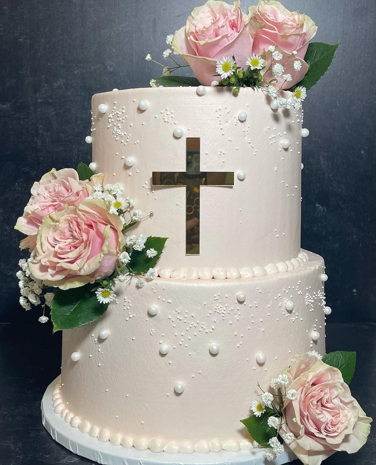 Two-tiered pink cake with gold cross and rose decorations for a religious celebration.