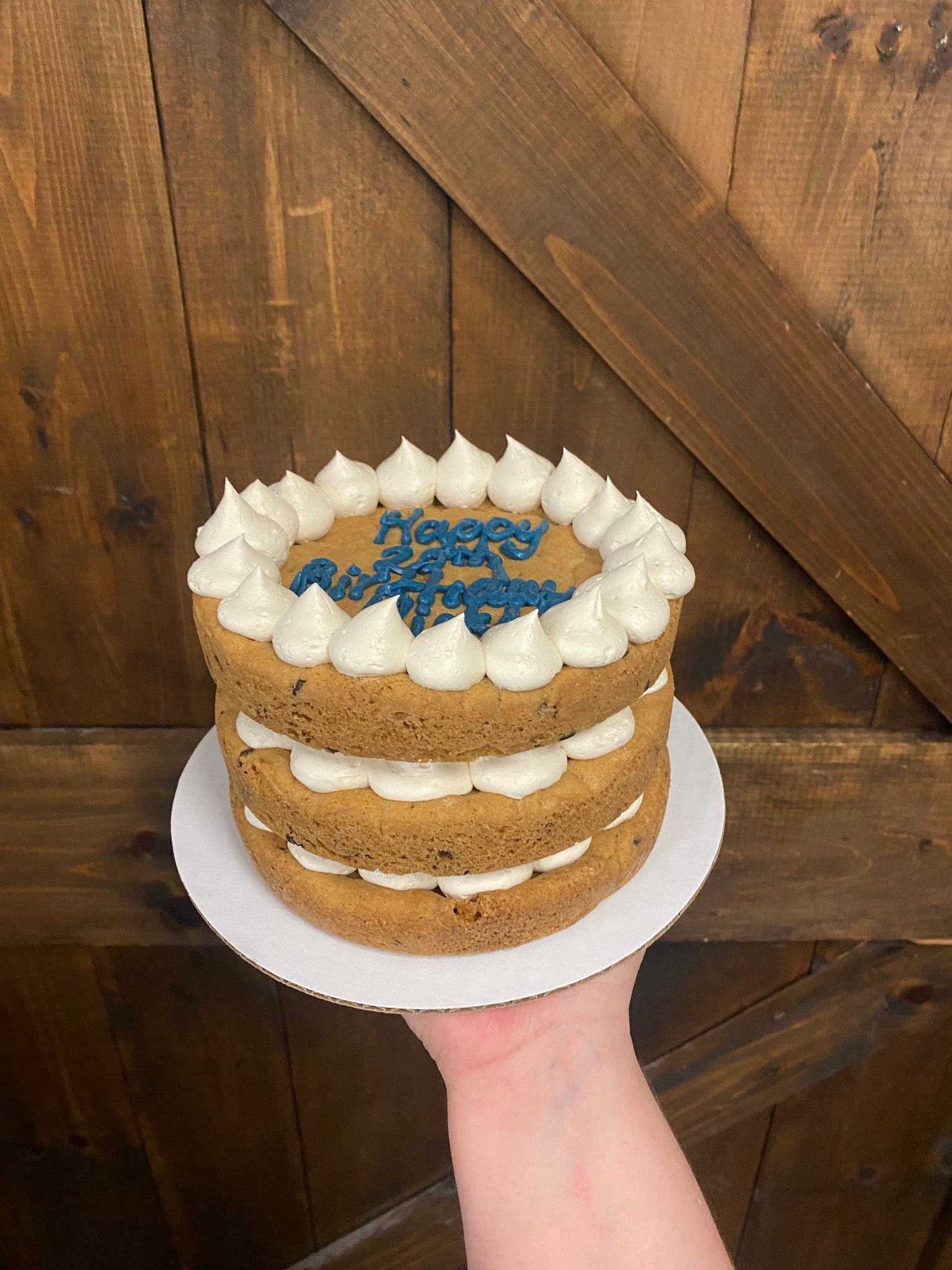 Three-layer cake with white frosting border and blue lettering, held in front of a wooden door.