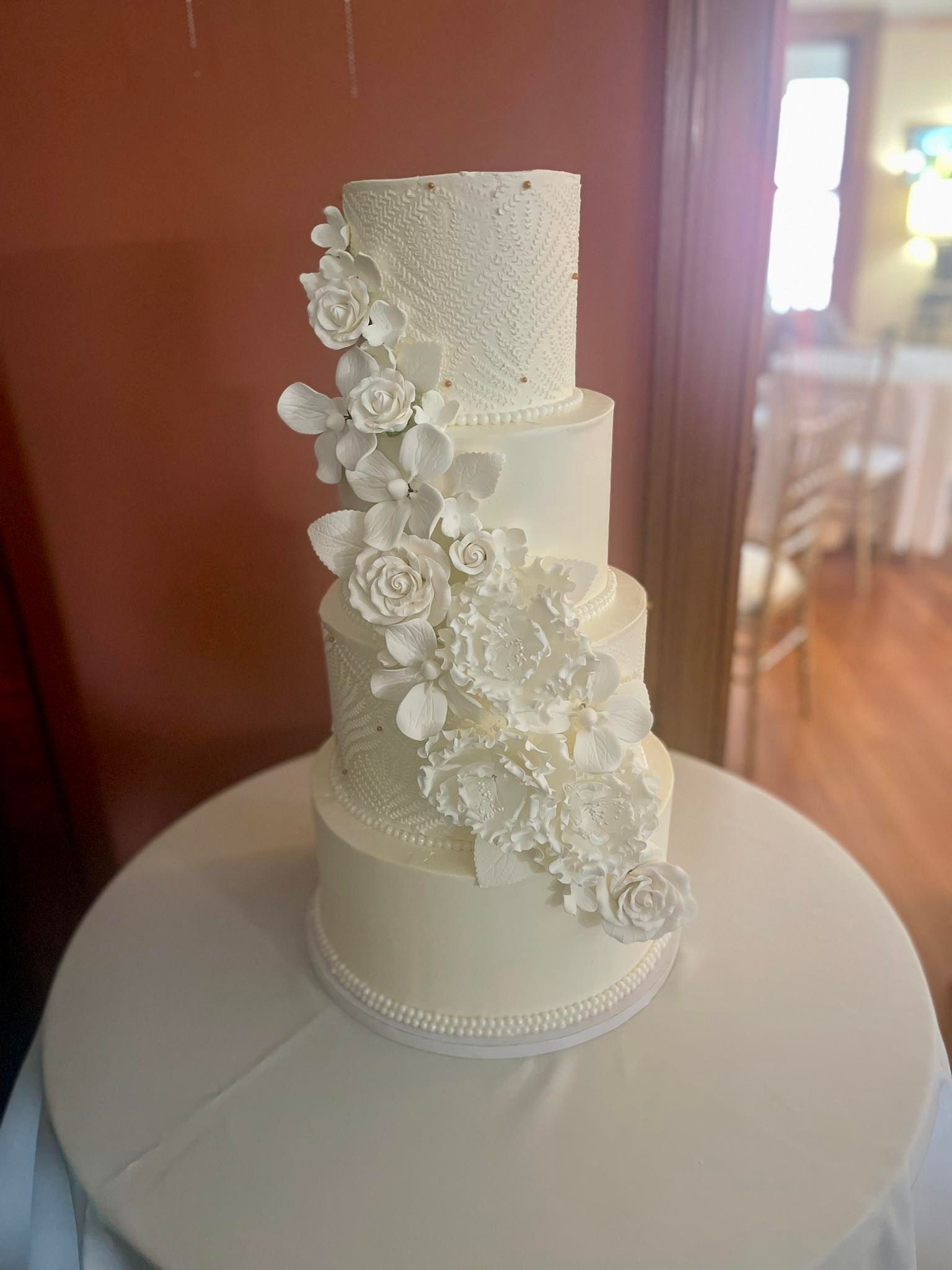 Four-tiered white wedding cake with cascading sugar flowers, on a white tablecloth, indoors.