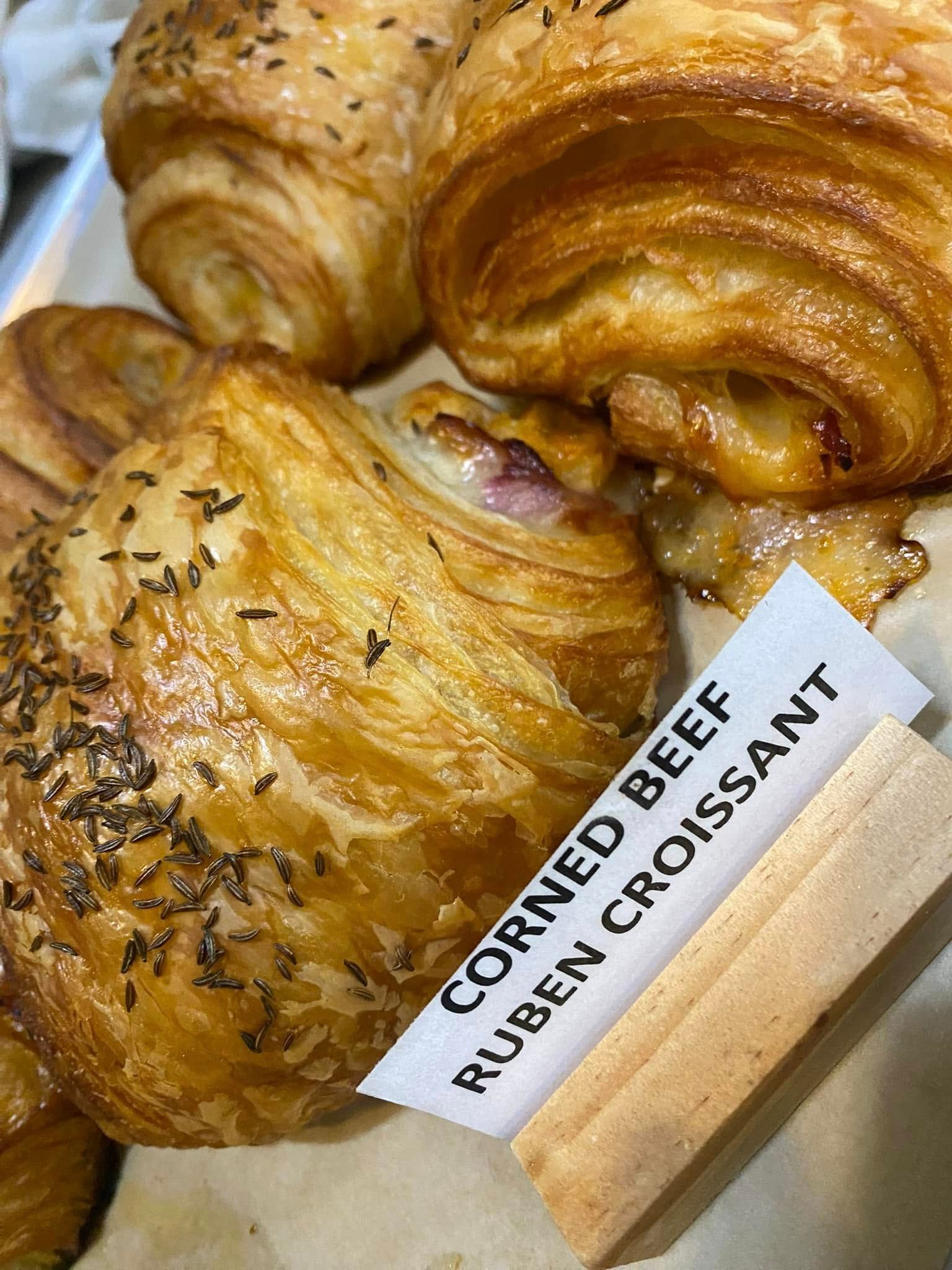 Close-up of golden, flaky corned beef Reuben croissant, topped with seeds, and a label.