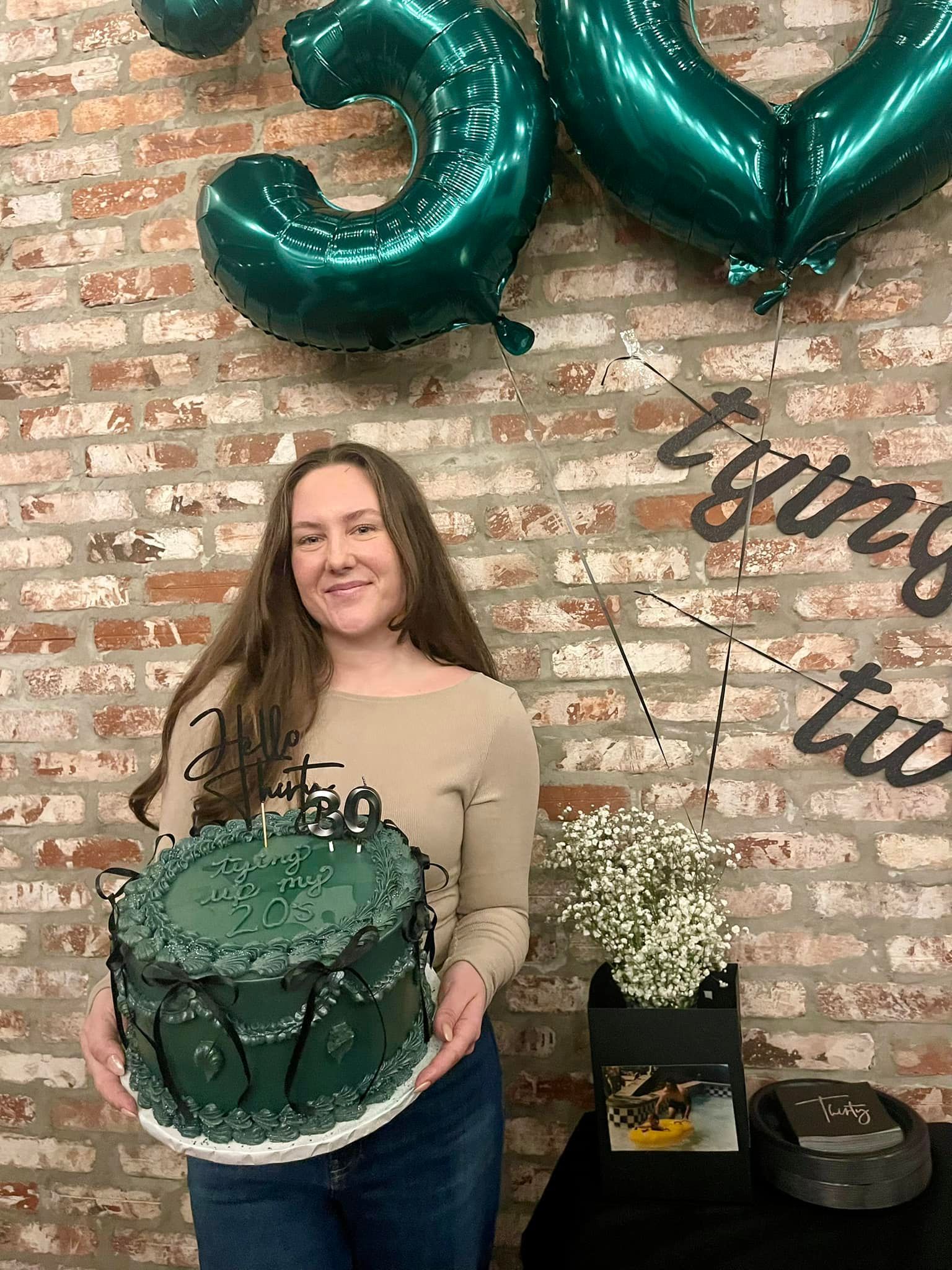 Woman holding a green cake, celebrating her 30th birthday with balloons and decorations in front of a brick wall.