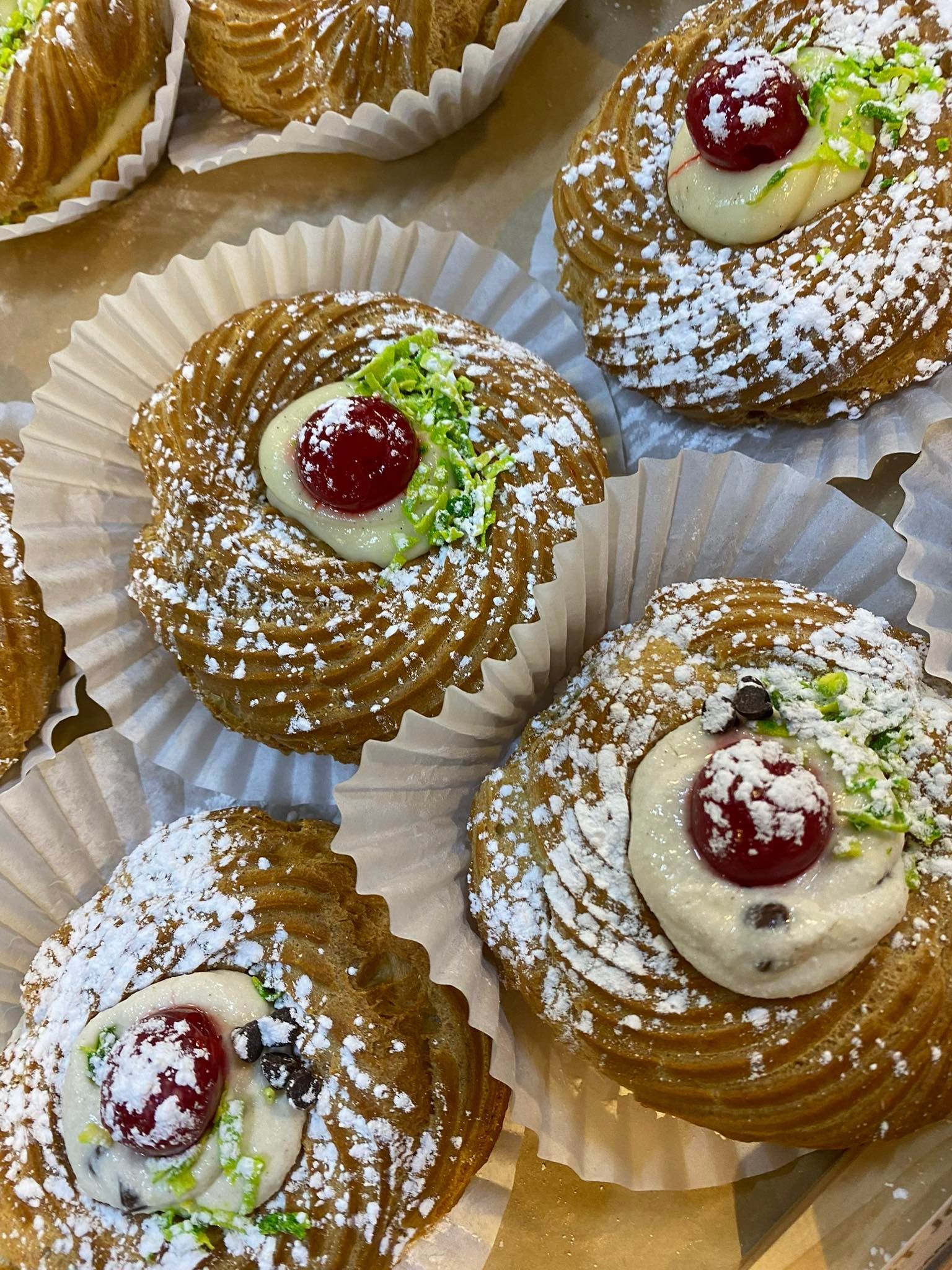 Close-up of pastries in paper cups, dusted with powdered sugar, topped with cream, cherry, and green garnish.