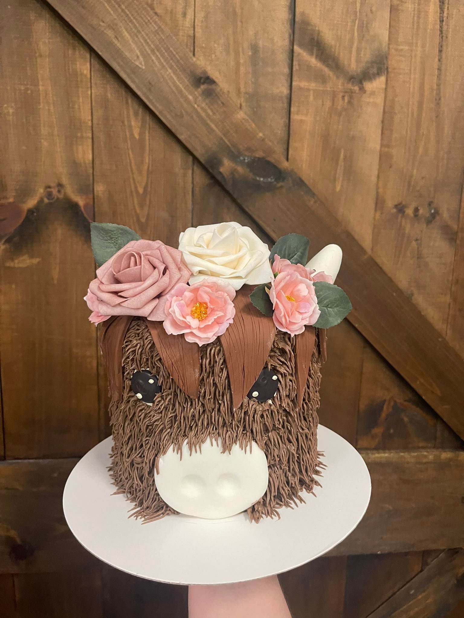 Cake decorated to look like a highland cow with chocolate fur, floral crown, and a white, fondant nose, set against a wooden door.