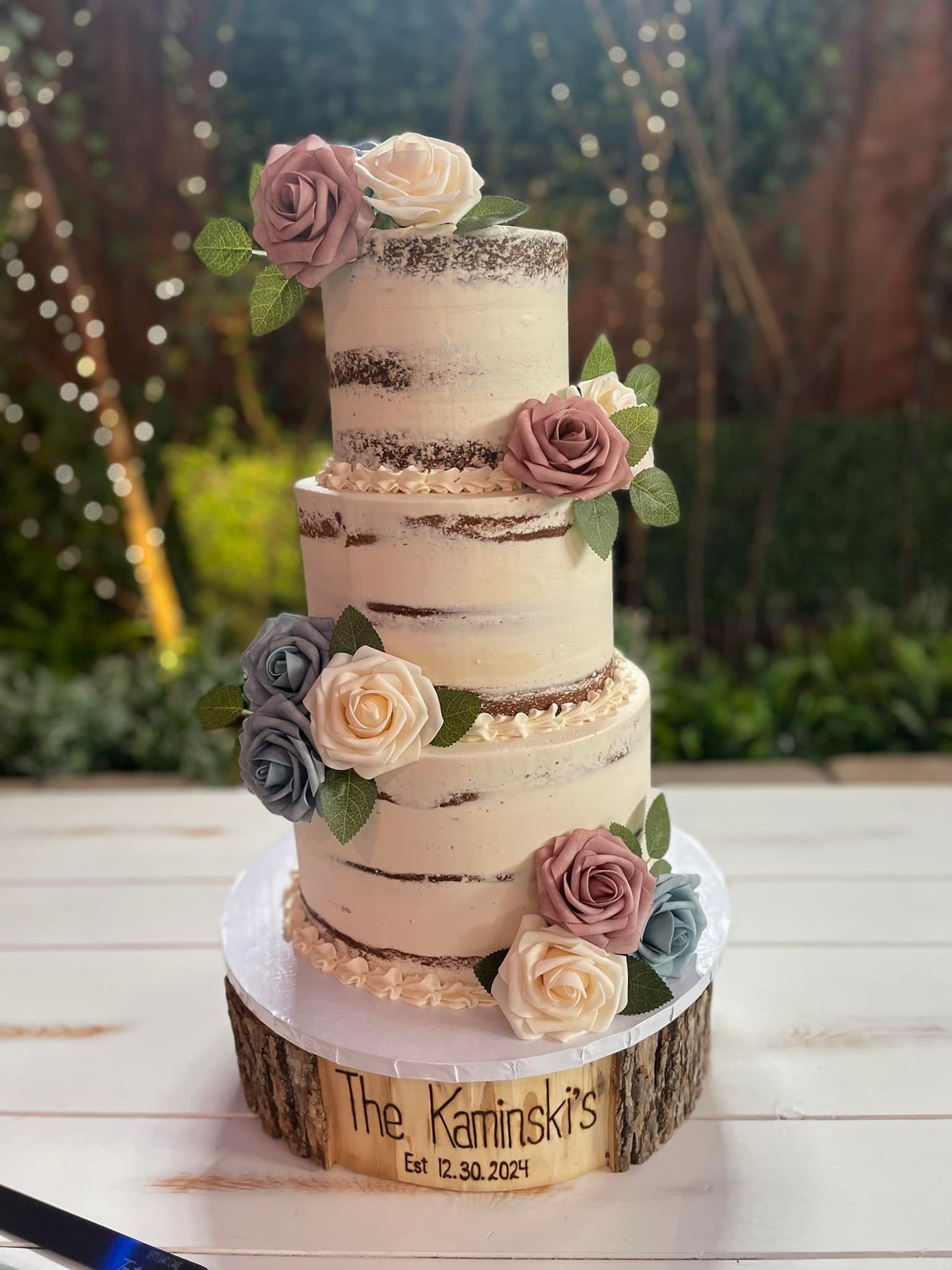 Three-tiered naked cake with floral decorations on a wooden cake stand outdoors.