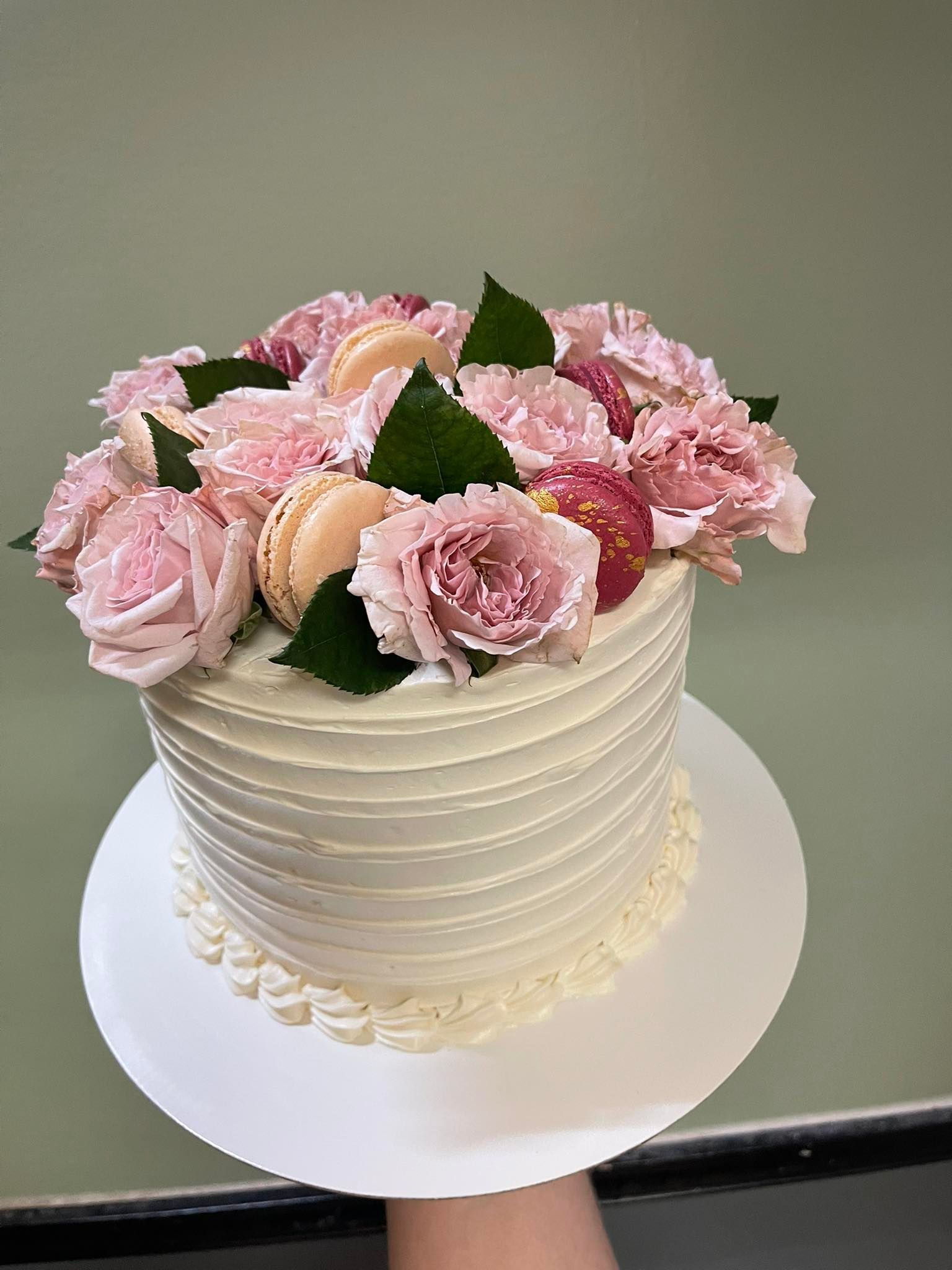 White cake decorated with pink flowers and macarons on a white cake board.