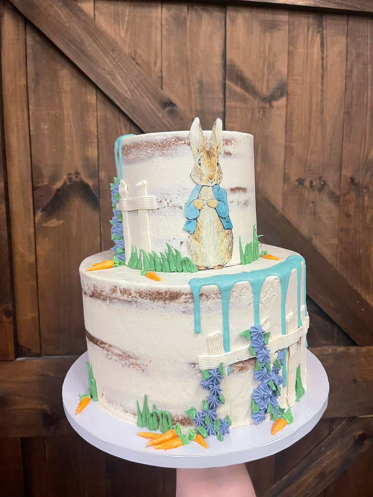 Two-tiered cake with blue drip, bunny topper, and decorative carrots and flowers, on a white plate in front of wood.