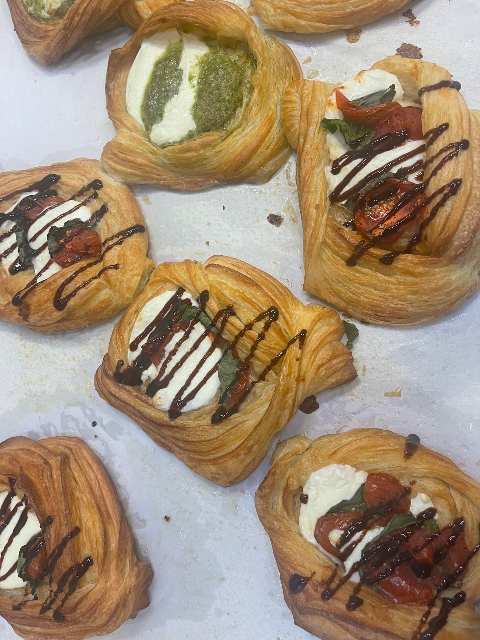 Assorted baked pastries topped with pesto, mozzarella, and balsamic glaze on parchment paper.