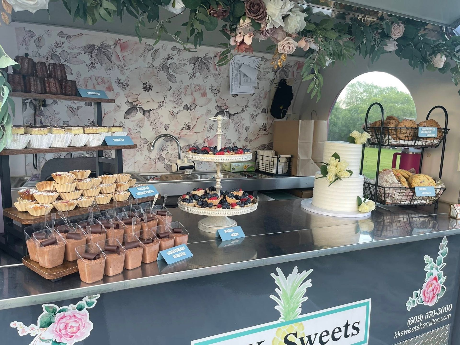 Food stall with pastries, tiered fruit displays, and a cake. Silver counter, floral decor, and a cart with a sign.