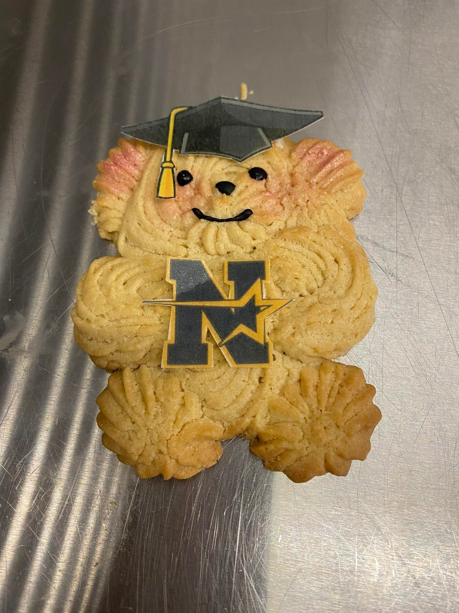 Cookie in the shape of a bear wearing a graduation cap, holding a logo with the letter 