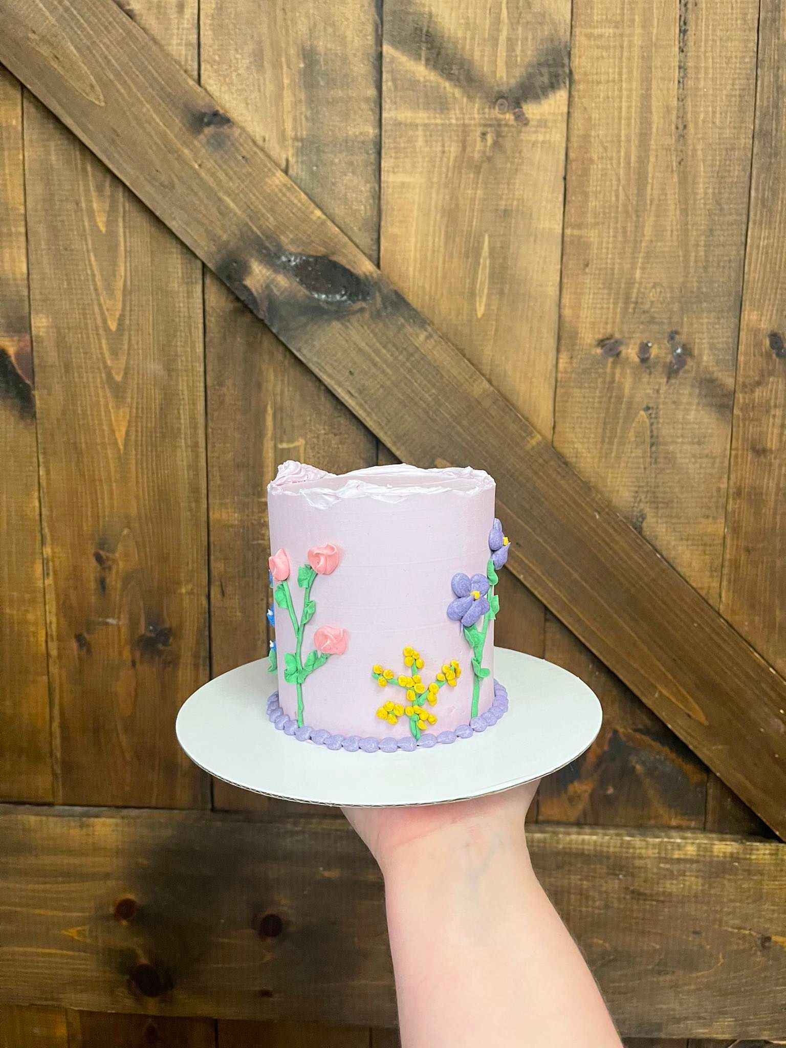 Hand holding a small lavender cake decorated with buttercream flowers on a white plate, against a wooden door.