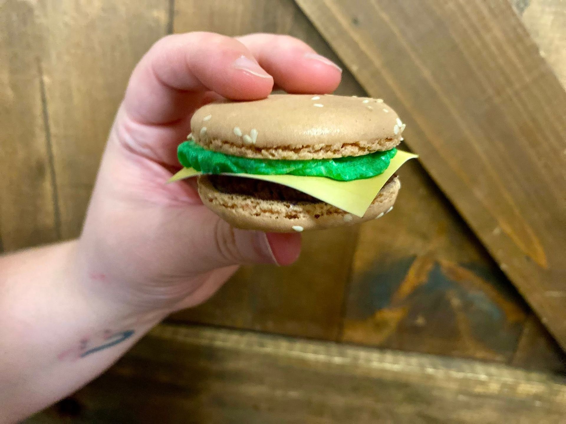 Hand holding a macaron designed like a mini burger; brown shell 